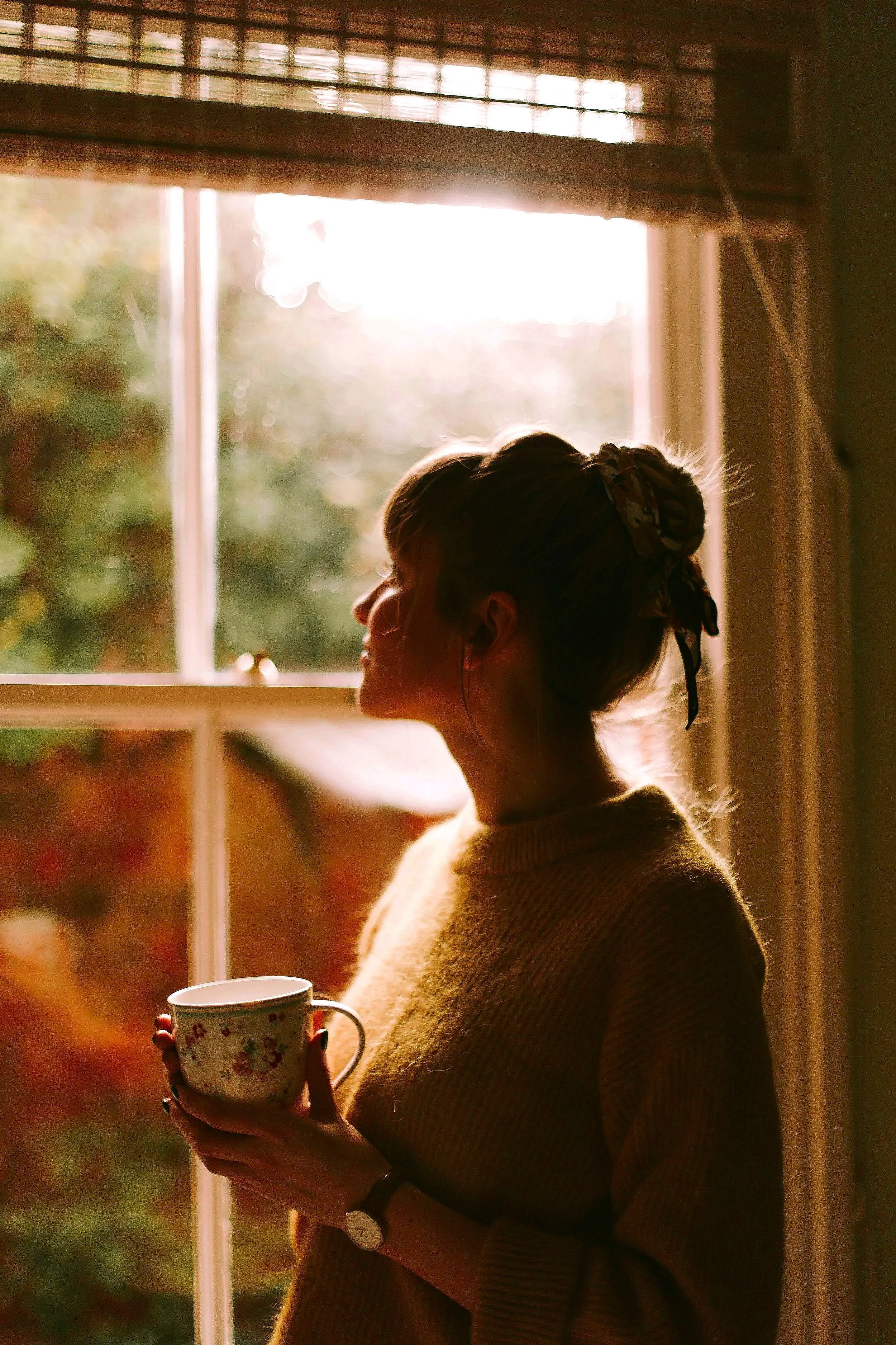 A woman holding a floral mug, standing by a window with sunlight shining through, creating a silhouette.