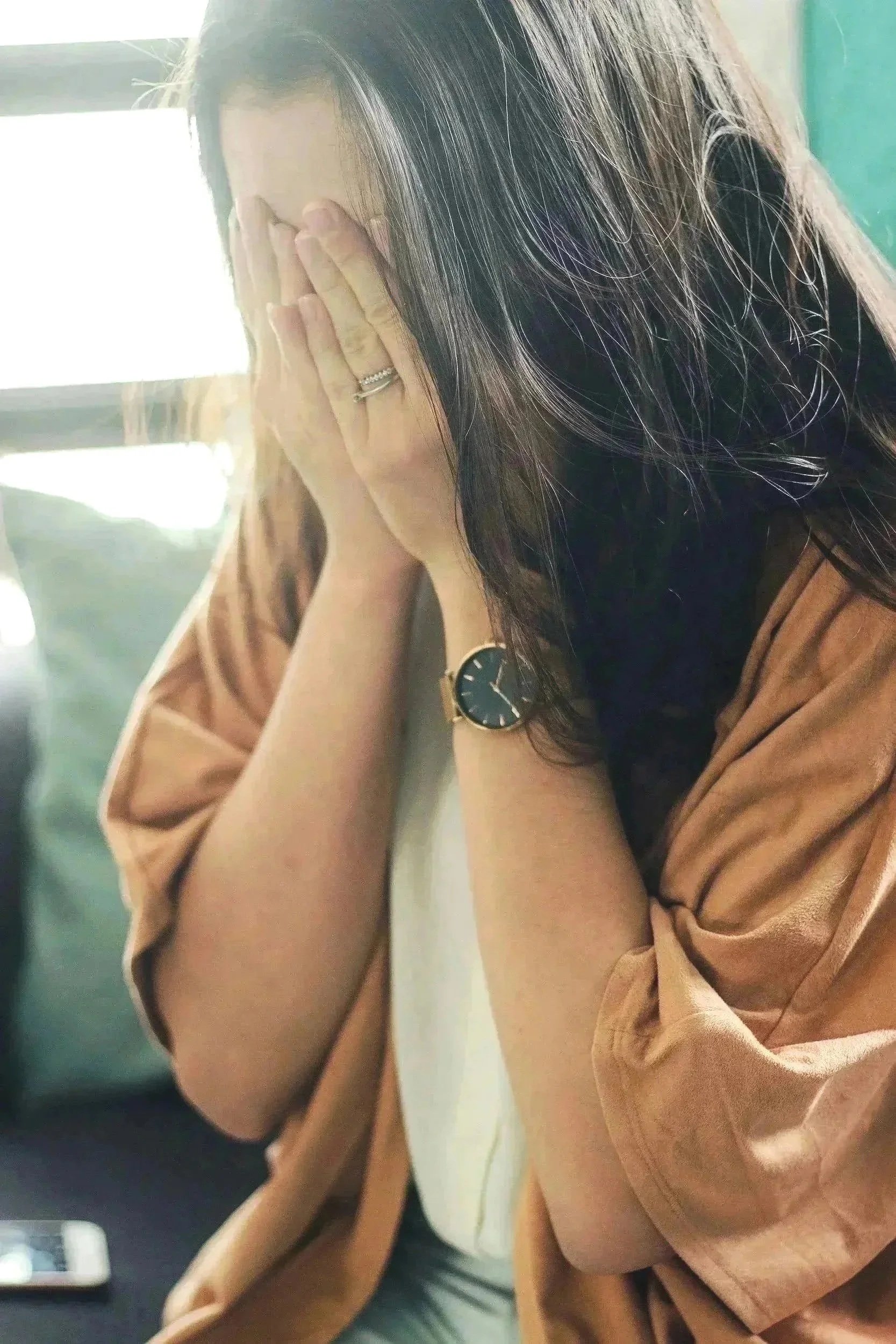 A woman with dark hair, wearing a watch and rings, sitting with her head in her hands.