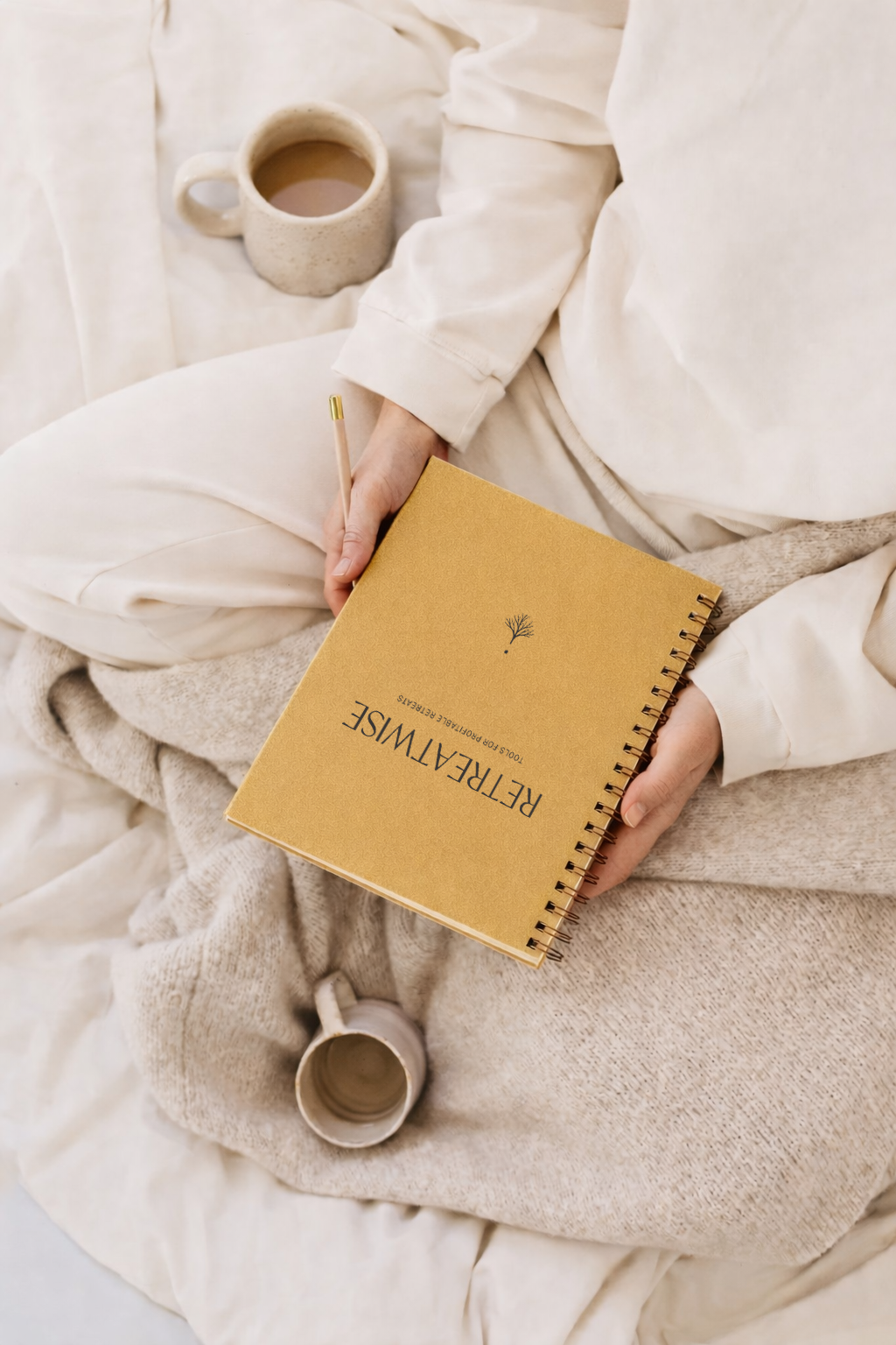 Person sitting cross-legged on bed, holding a tan hardcover notebook with the word "REFLECTIONS" on the cover, a pencil, a beige mug, and a ceramic cup filled with coffee, all surrounded by cozy white and beige bedding.