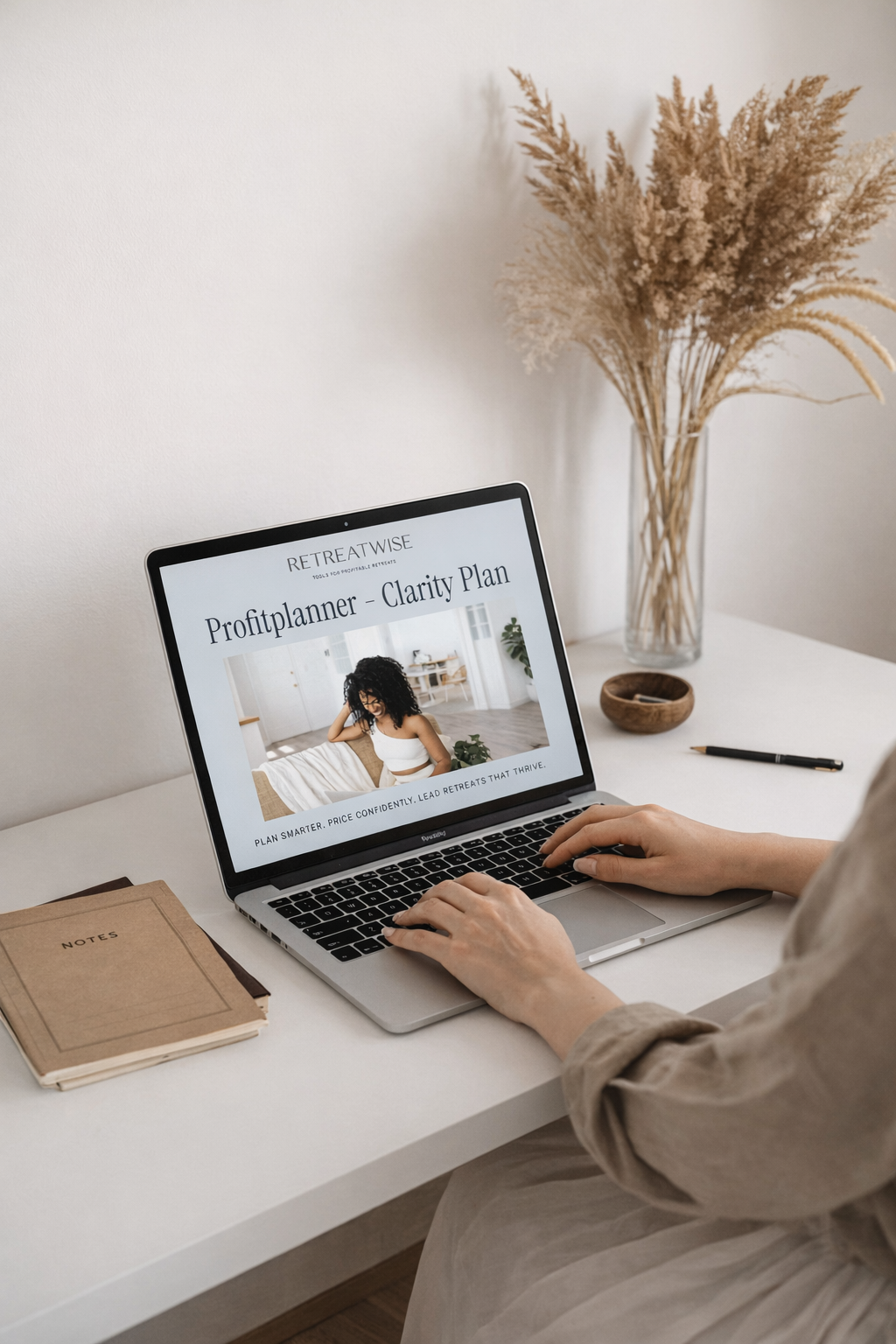 A person working on a laptop with a Profitplanner Clarity Plan webpage, on a white desk with a notebook, pen, and a vase of dried flowers in the background.