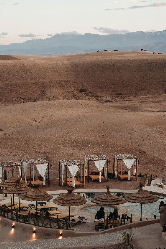 A desert resort with cabanas near a swimming pool, overlooking brown rolling hills and distant mountains.