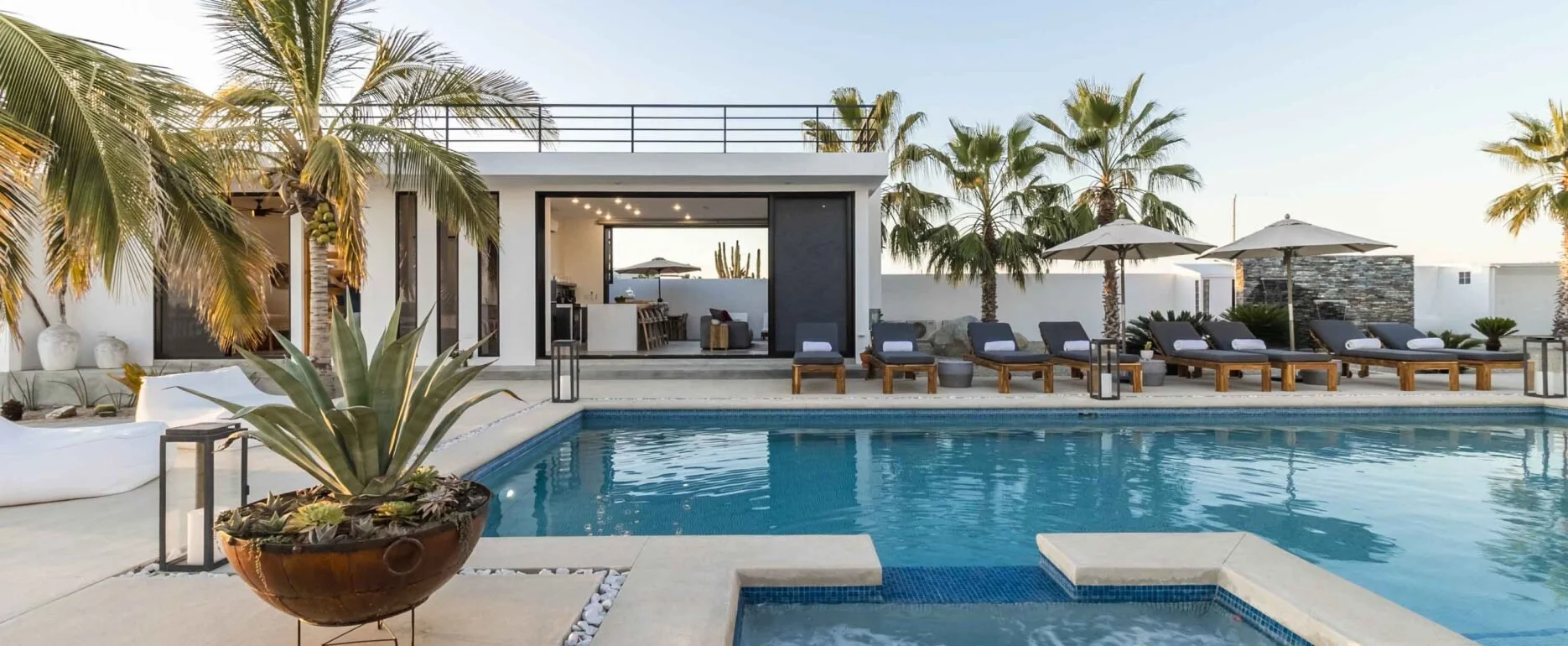 Modern backyard with a swimming pool, sun loungers, umbrellas, palm trees, and a house with large sliding doors open to an outdoor kitchen and seating area at sunset.