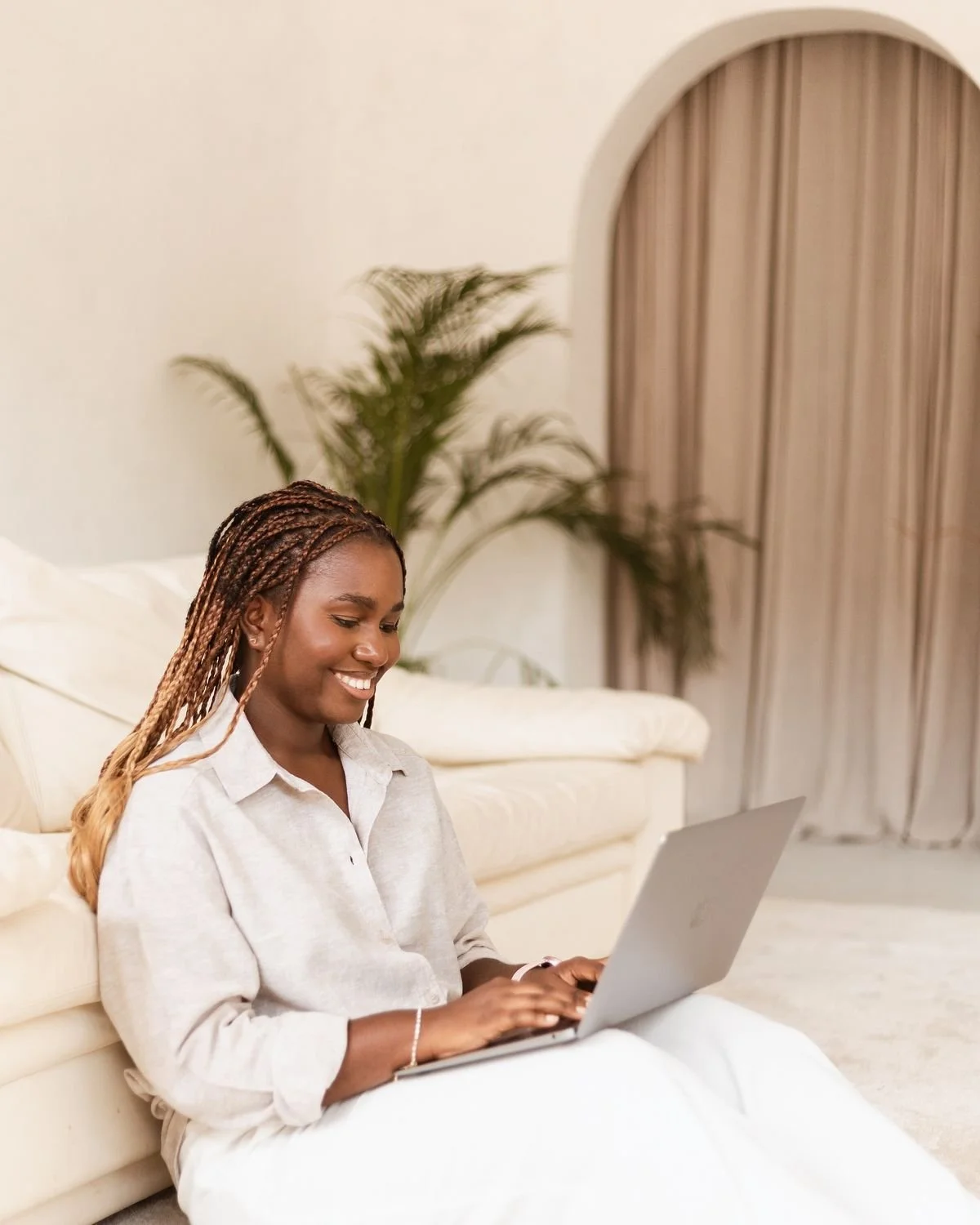 A woman with braided hair sitting on a white couch using a silver laptop in a living room with beige curtains and a green plant in the background.