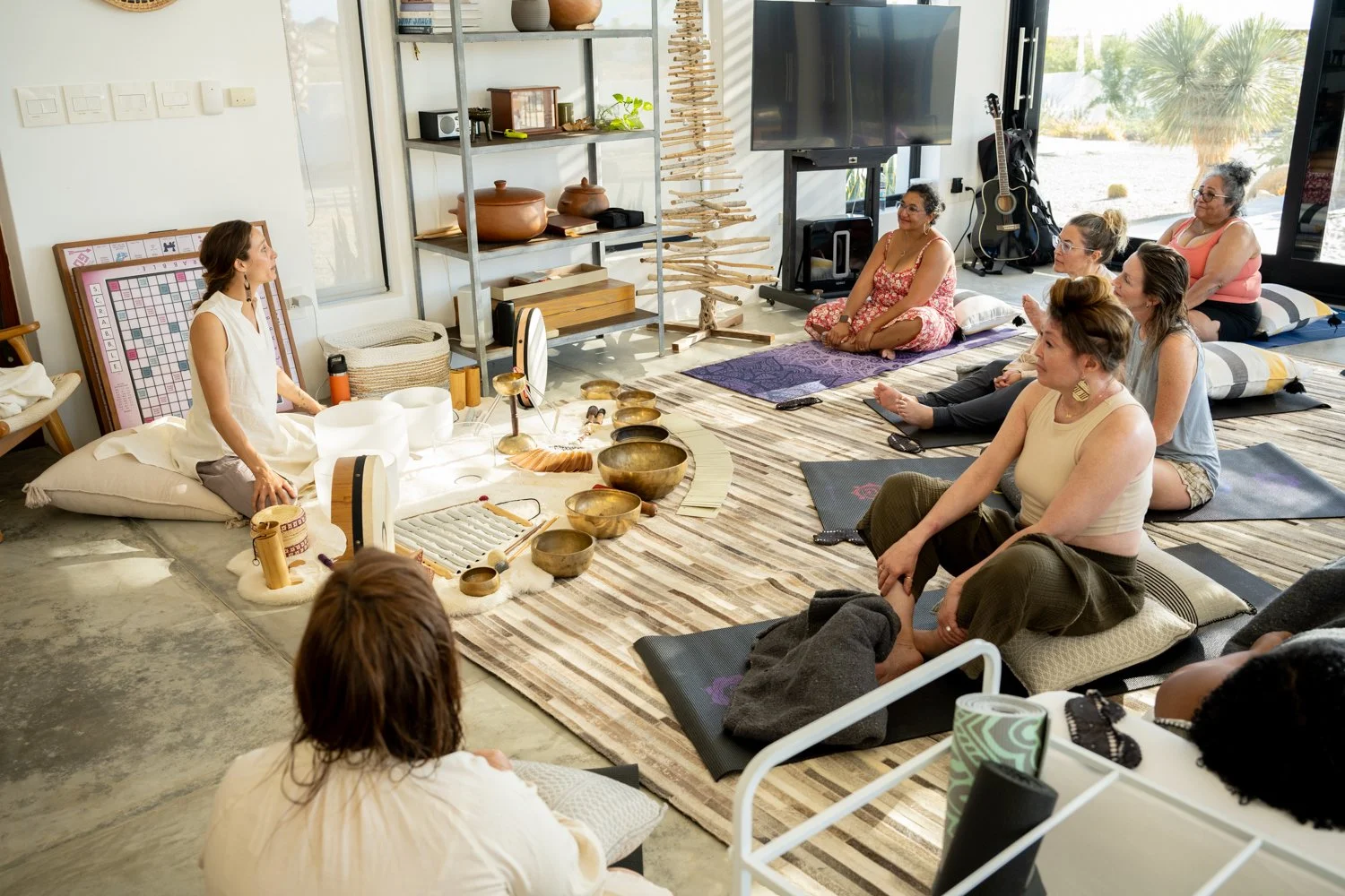 Women participating in a meditation or sound therapy session in a bright, modern room with natural light. The instructor sits on the floor surrounded by singing bowls and a white drum, with participants sitting on yoga mats or cushions listening attentively.