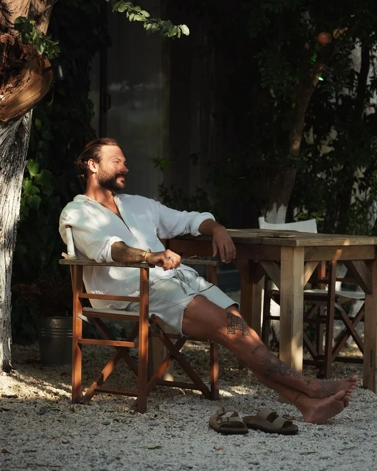 A man with tattoos and a beard sitting relaxed outdoors under a tree, wearing a white shirt and shorts, with sandals on the ground in front of him, smiling and enjoying the moment.