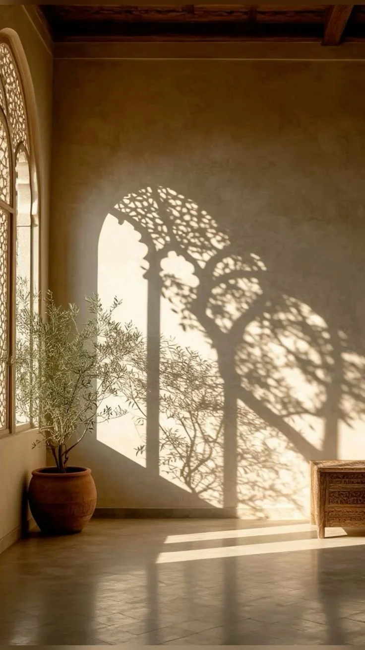 Sunlight casts the shadow of a decorative, ornate window grille onto an interior wall, creating a detailed pattern in a room with beige walls, wooden ceiling beams, a potted plant, and a small wooden bench.