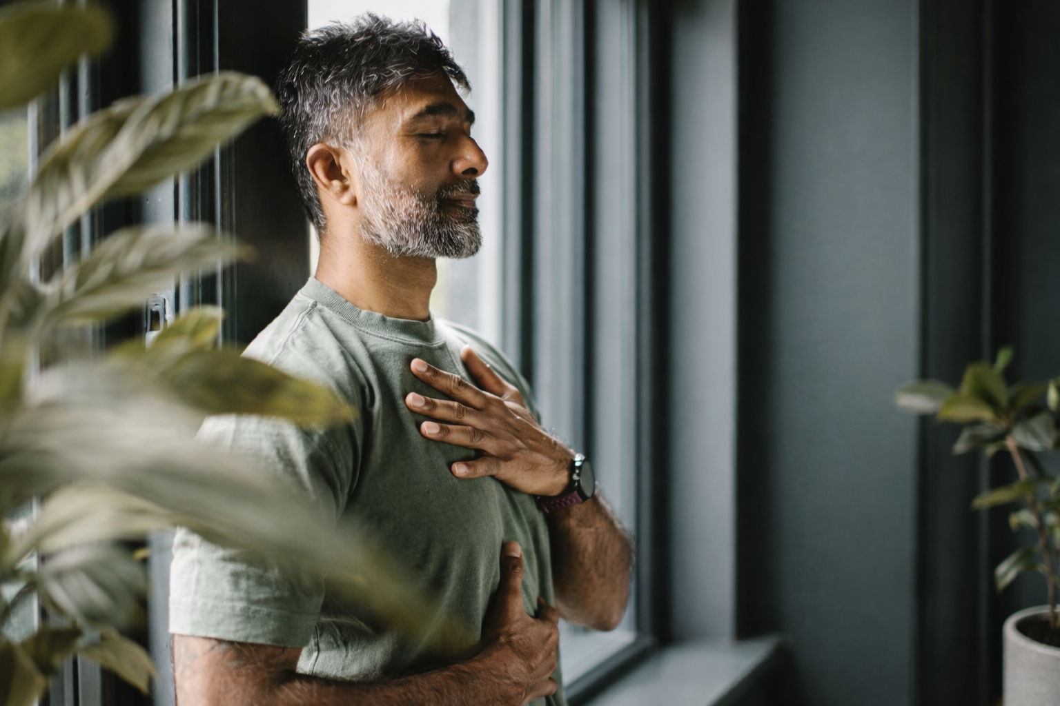A man with gray hair and a beard practicing meditation with eyes closed, hand on chest, in a room with large windows and plants.