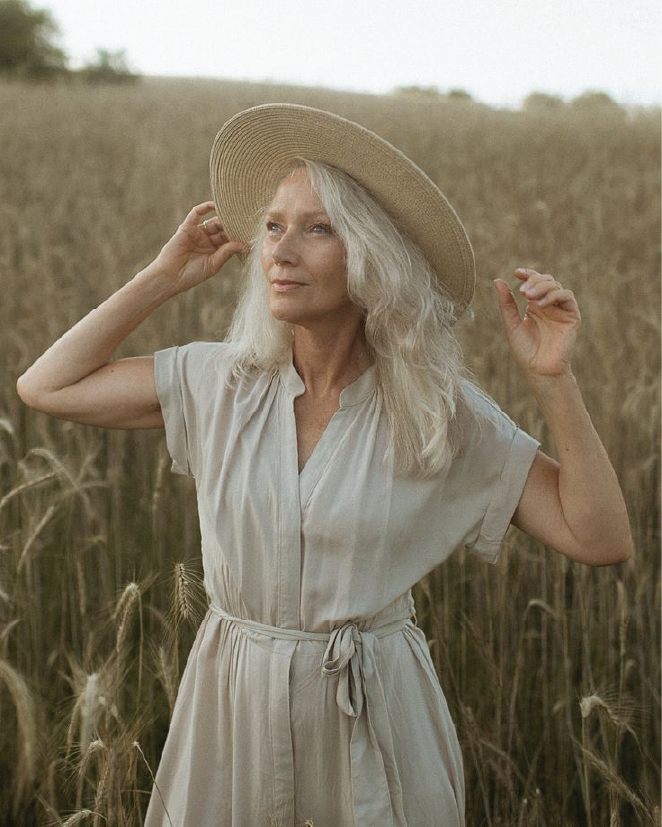 An elderly woman with long gray hair standing in a field of wheat, wearing a beige dress and a wide-brimmed straw hat, touching her hat with both hands and looking into the distance.