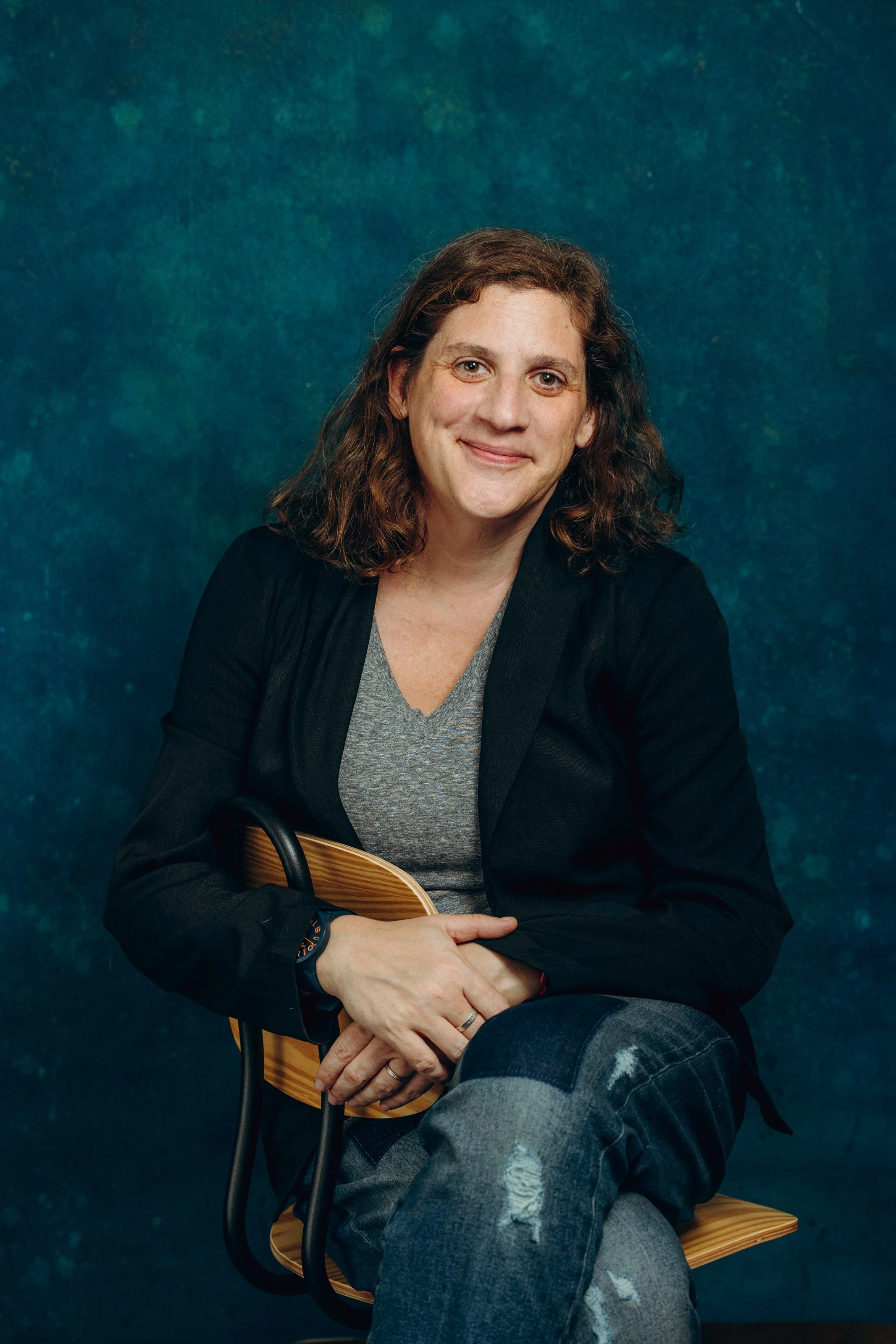 A woman with shoulder-length curly brown hair sitting on a wooden chair against a blue textured background, wearing a black blazer, gray t-shirt, and ripped jeans, holding her hands on her lap and smiling at the camera.