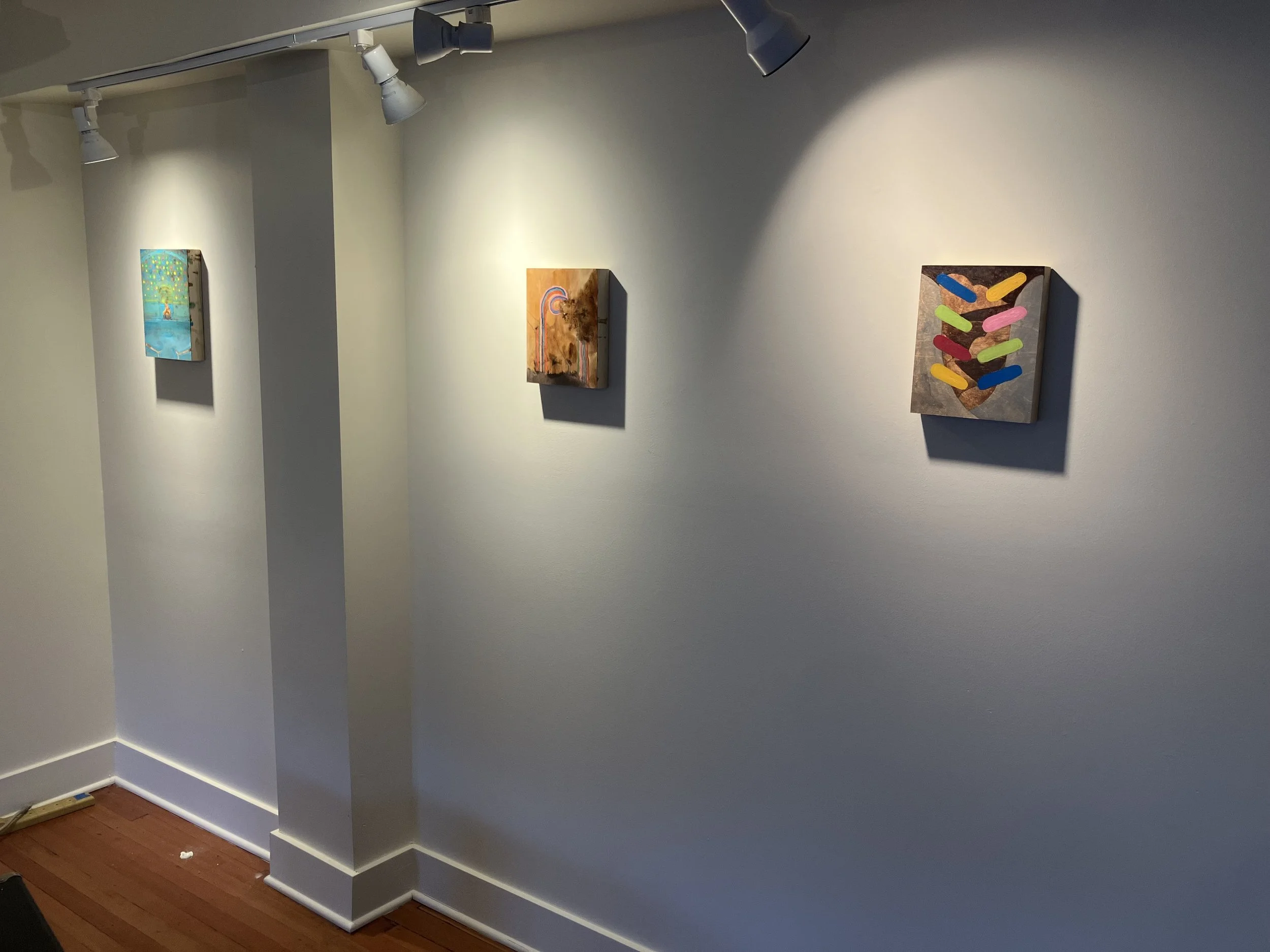 White gallery wall with three small, colorful paintings.