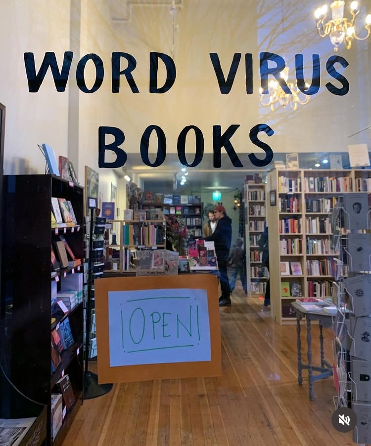 Front window of bookstore with Word Virus Books written on glass in black and an "Open" sign below. Bookshop has multiple bookshelves and two men standing at the counter.