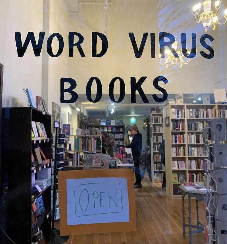 Photo of a bookshop through a glass window with the words "Word Virus Books" written in black and an Open sign below. Bookshop has two people standing at the counter.