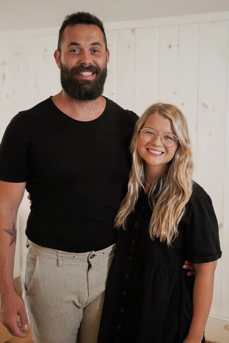 Un homme et une femme souriants posent ensemble dans une pièce lumineuse. L'homme porte un t-shirt noir et un pantalon beige à carreaux, la femme porte une robe noire avec des lunettes.