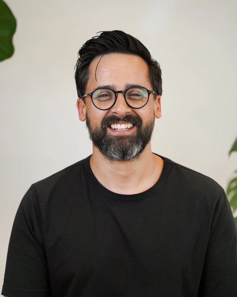 Homme souriant avec lunettes, barbe, cheveux noirs, portant un t-shirt noir, fond neutre avec une plante verte sur le côté gauche.