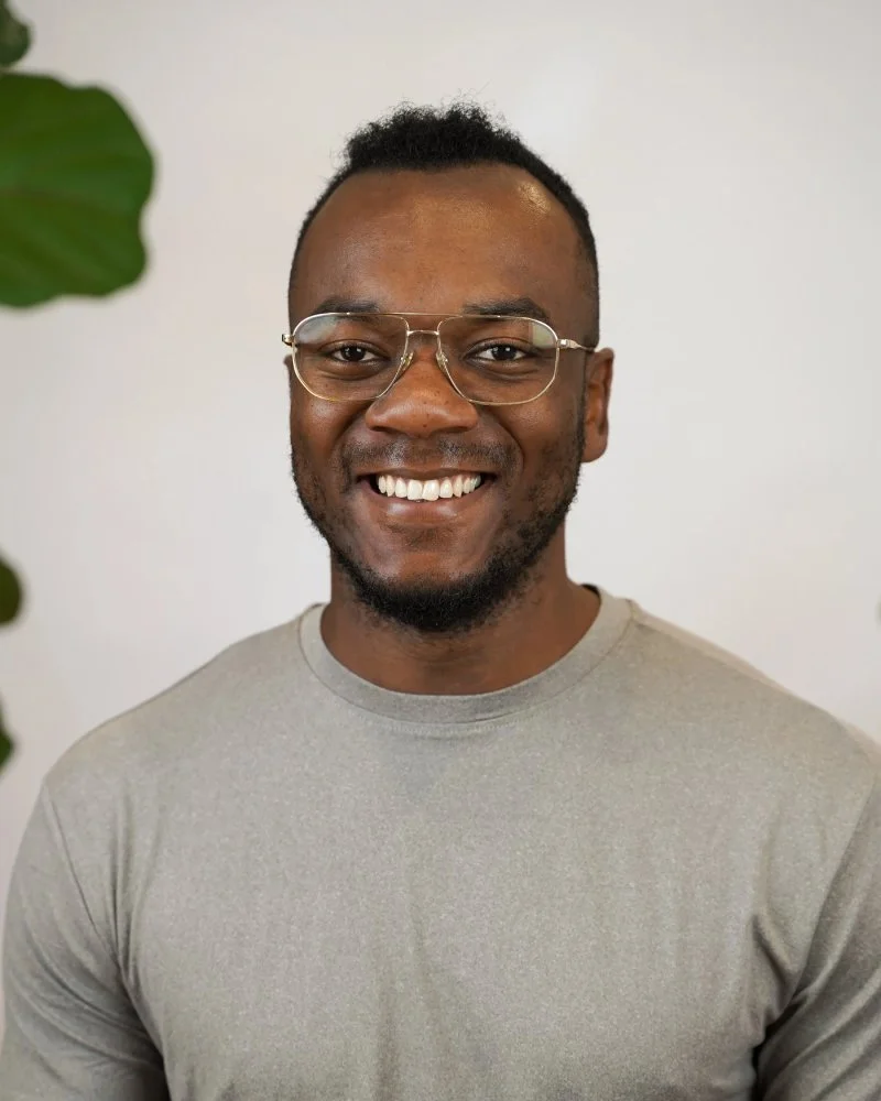 Homme souriant portant des lunettes, fond blanc, partie d'une plante verte visible à gauche.