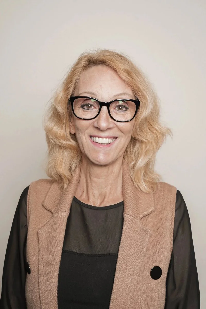 Femme blonde avec des lunettes noires, souriante, portant un haut noir et un blazer beige, debout devant un mur clair.
