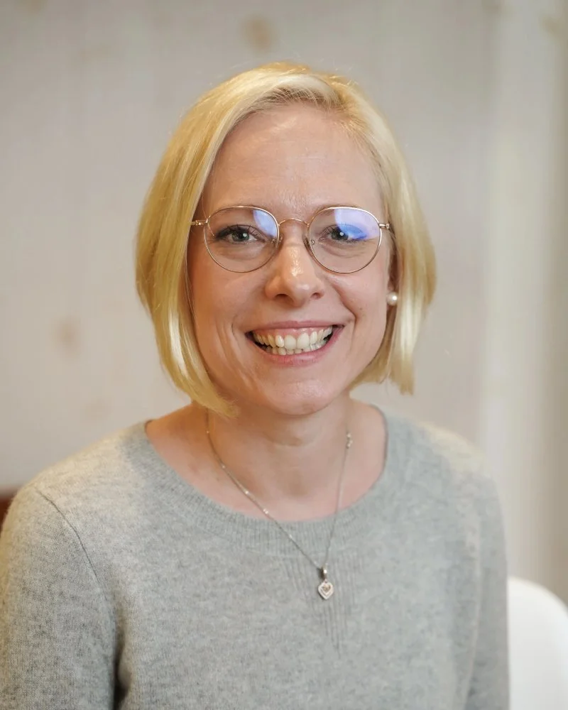 Femme blonde souriante portant des lunettes rondes, un pull gris, une paire de boucles d'oreilles perles et un collier avec un pendentif en forme de cœur.