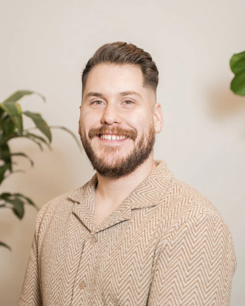 Portrait d'un homme souriant avec une barbe, portant une chemise beige à motif herringbone, arrière-plan avec des plantes vertes.