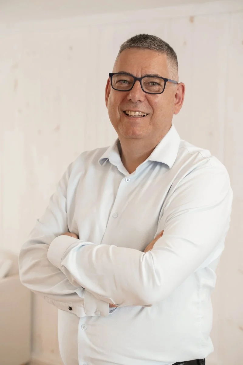 Un homme souriant avec des lunettes, habillé en chemise blanche, se tient les bras croisés