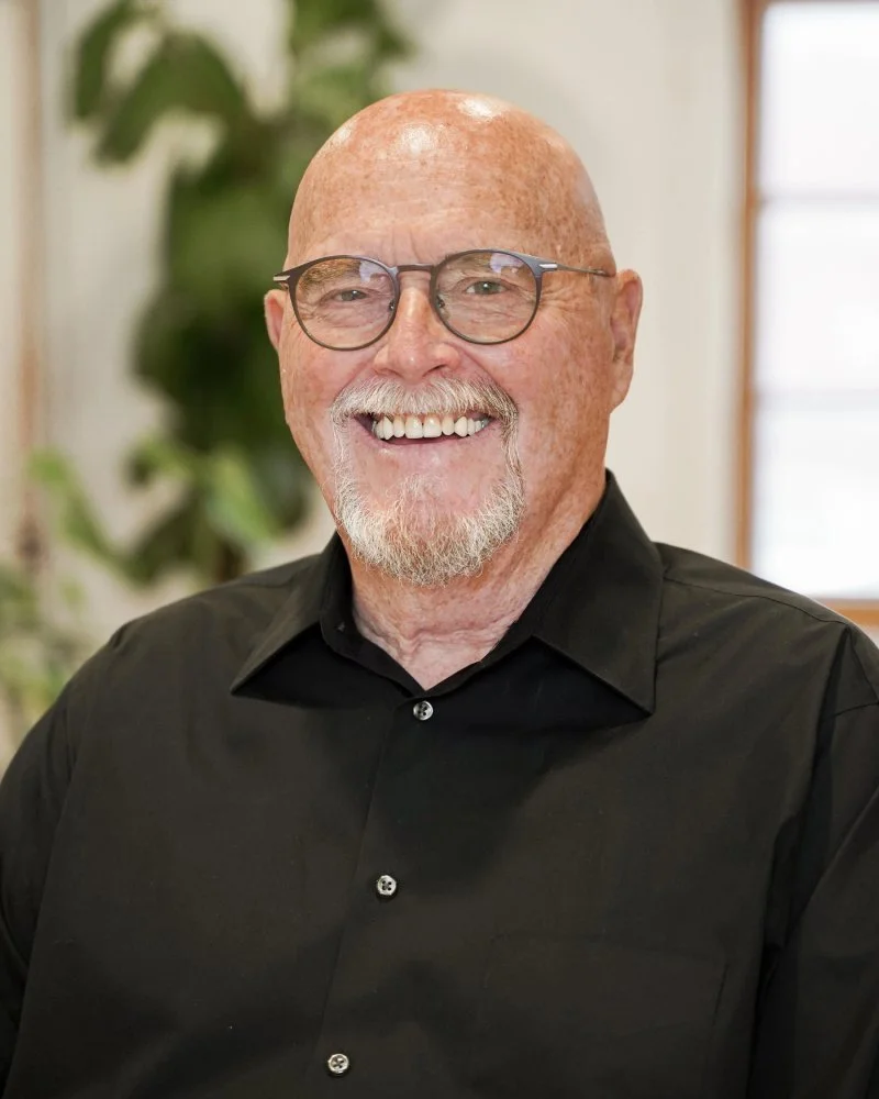 Portrait d'un homme âgé souriant portant des lunettes et une chemise noire, avec un fond flou de plantes et de fenêtres.