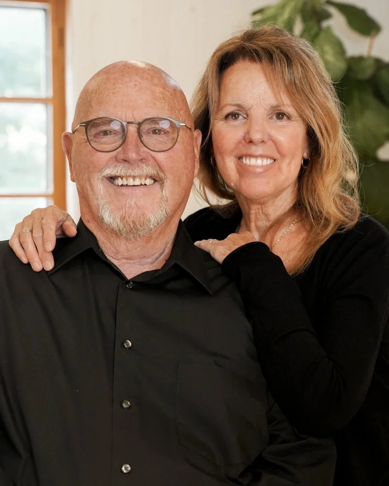 Un couple souriant pose ensemble dans une pièce lumineuse avec une fenêtre en arrière-plan et une plante verte à droite.