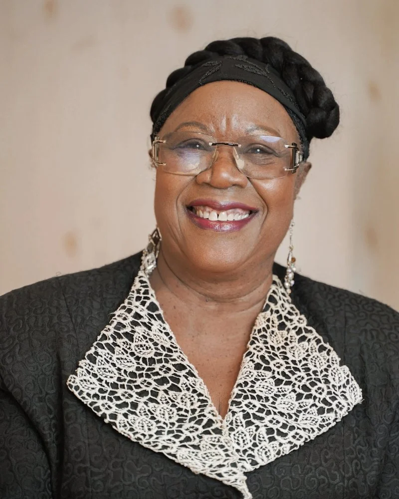 Une femme âgée souriante portant des lunettes, des boucles d'oreilles pendantes, une écharpe en dentelle blanche, et un vêtement noir avec un motif texturé, avec un fond neutre.