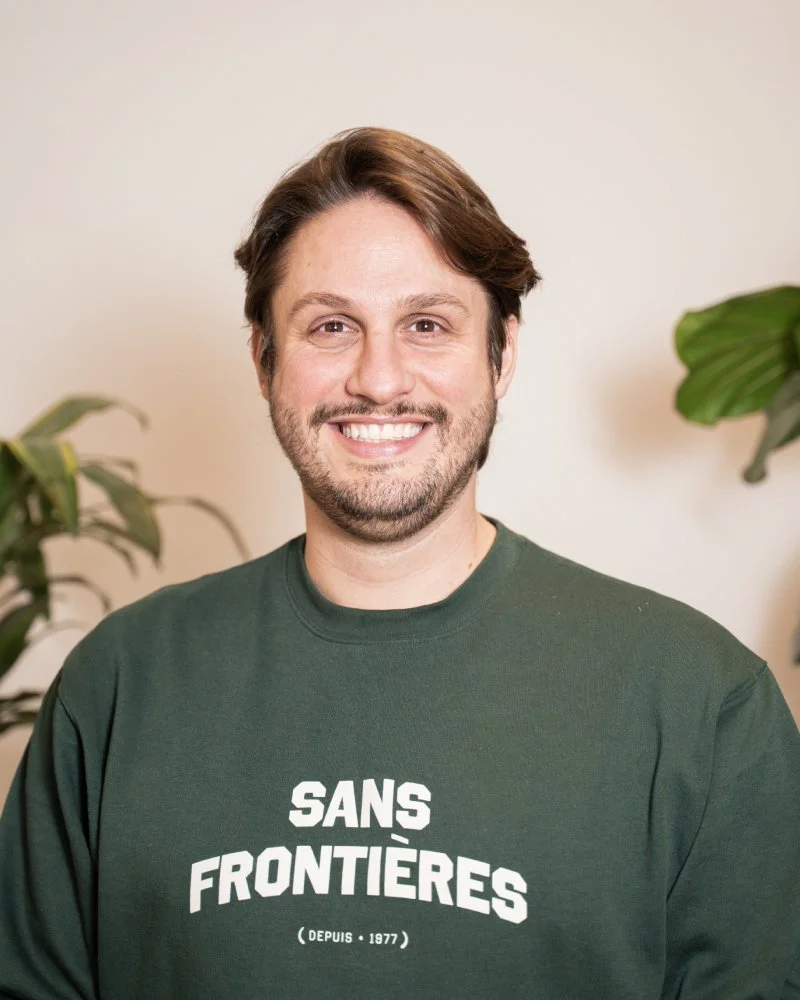 Un homme souriant avec des cheveux bruns et une barbe, portant un sweat vert avec l'inscription 'Sans Frontières'.
