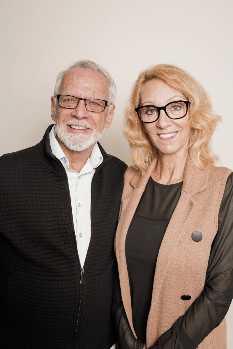 Un homme âgé et une femme portent des lunettes et sourient, debout côte à côte contre un mur blanc.
