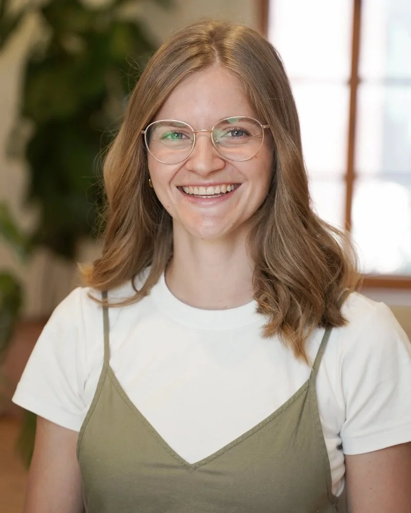 Jeune femme souriante portant des lunettes, une chemise blanche et une robe beige, dans un intérieur lumineux avec des fenêtres et des plantes en arrière-plan.