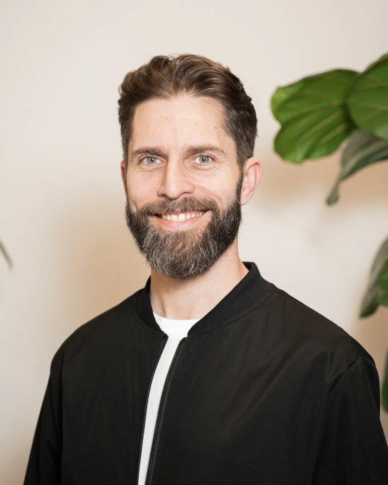 Un homme souriant avec une barbe, portant une veste noire, debout à côté de plantes vertes dans un décor intérieur.