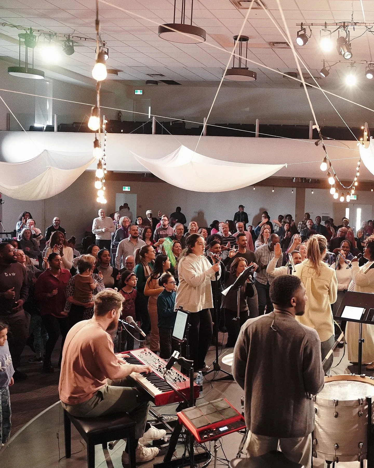 Un groupe de personnes assistent à un concert ou à une répétition musicale dans une salle voûtée avec des décorations légères suspendues.