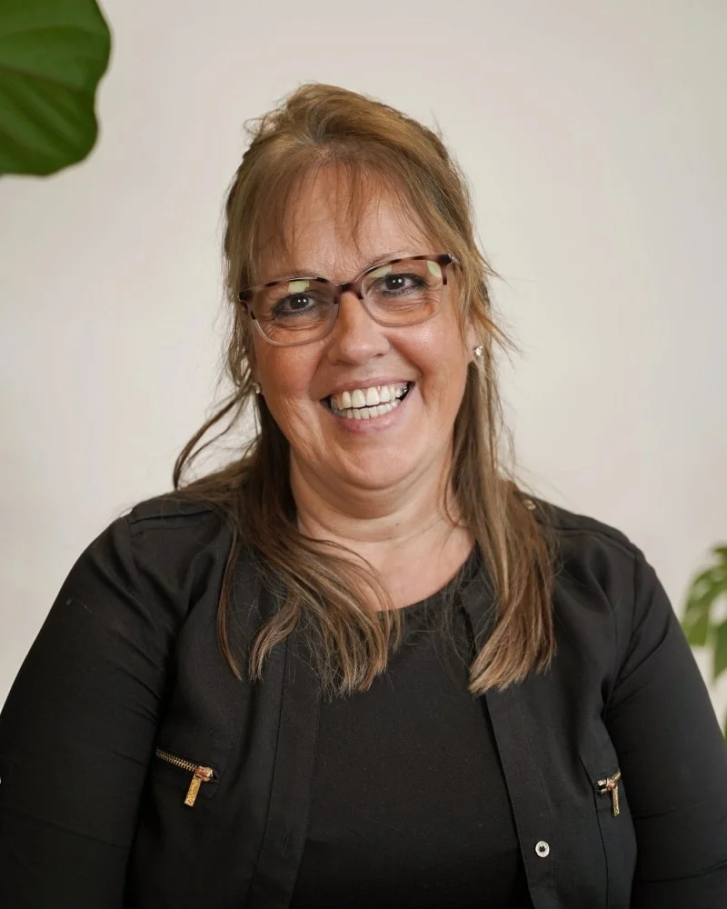 Une femme souriante avec des lunettes, portant un vêtement noir, face à la caméra, avec un fond clair et quelques plantes visibles.