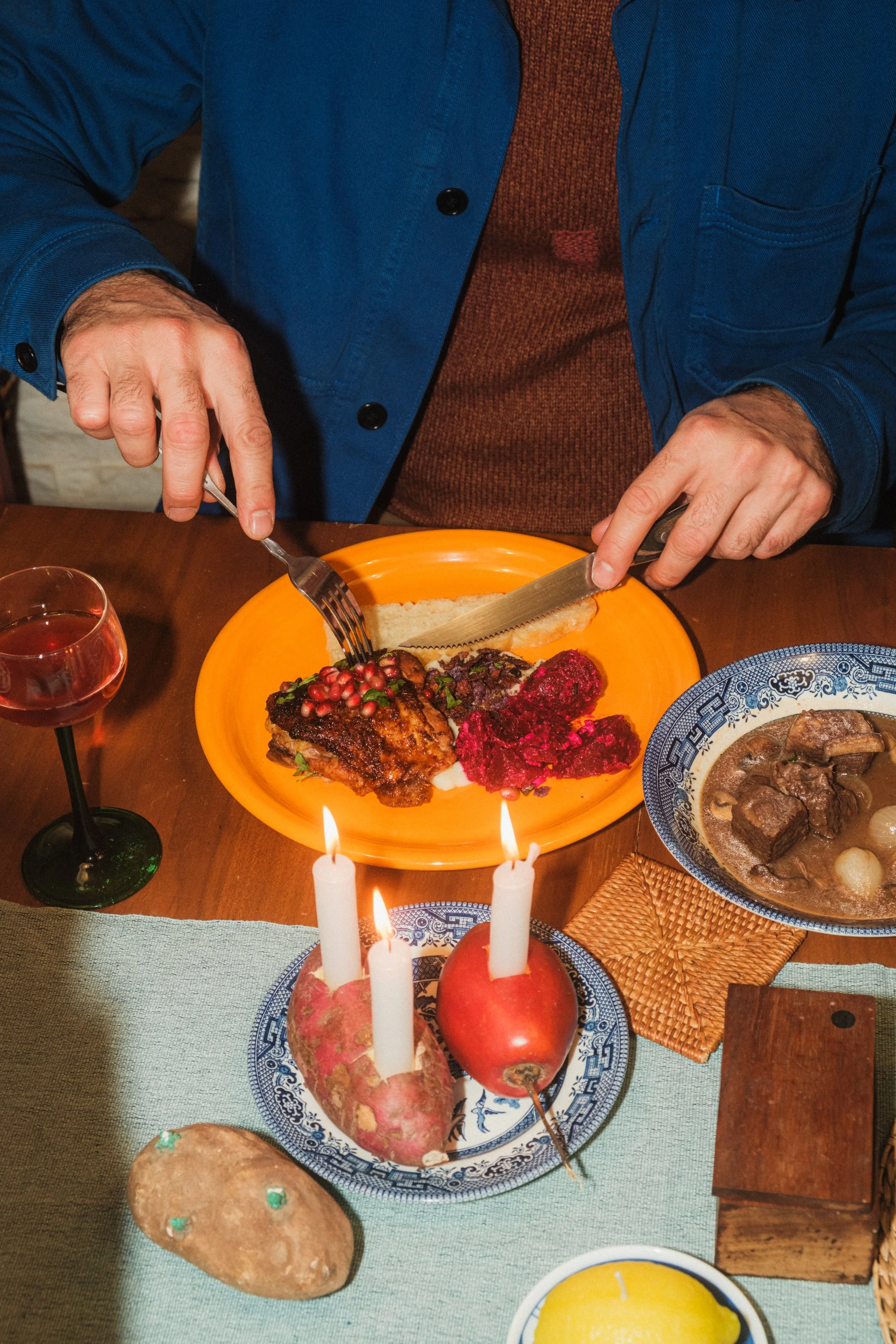 Man cutting food on an orange plate, surrounded by a glass of red wine, a bowl of beef stew, and festive candles on a table.