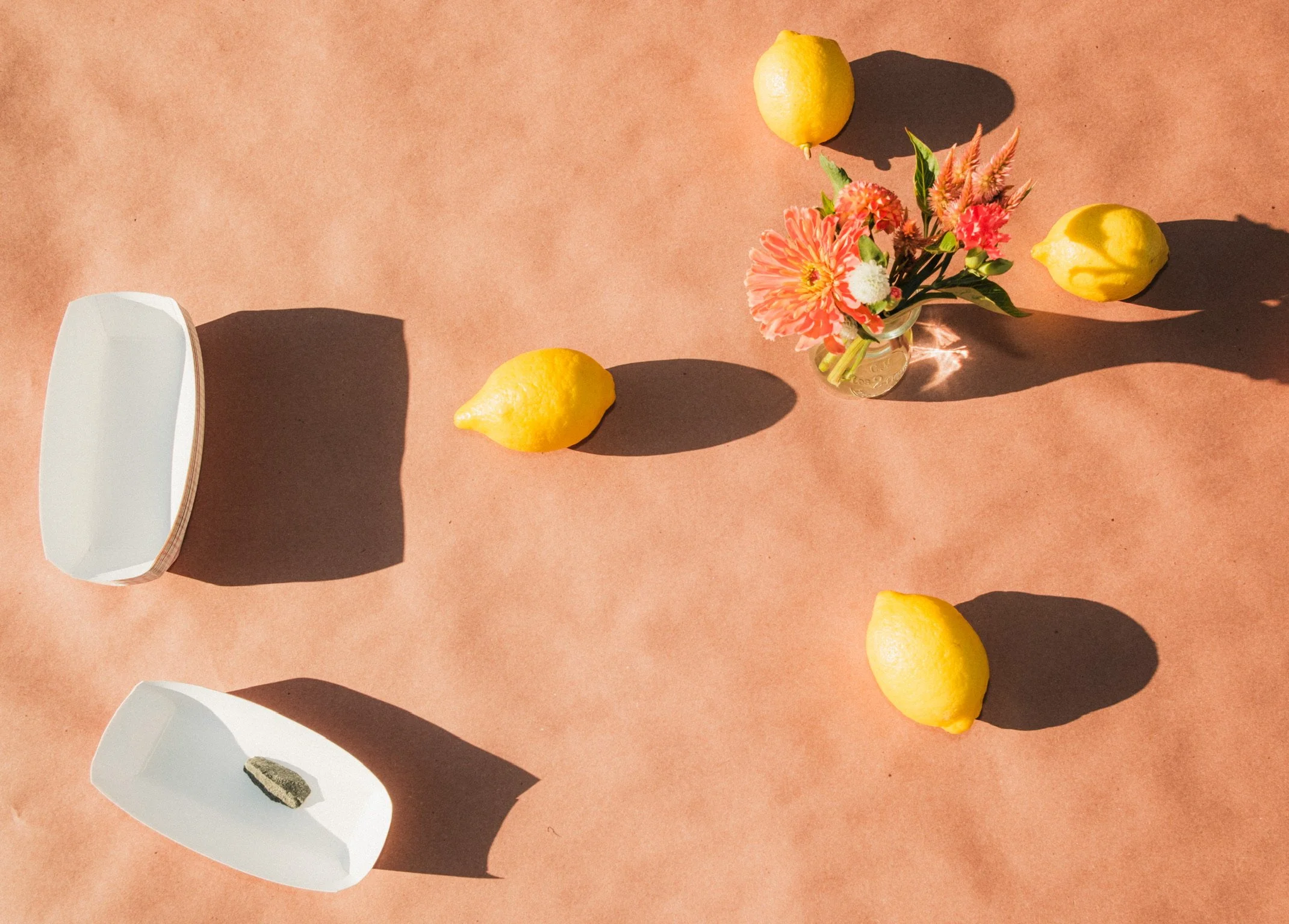 A flat lay of three lemons, a small vase with pink and orange flowers, and two paper hot dog boats with a stone object inside on a brown paper background.