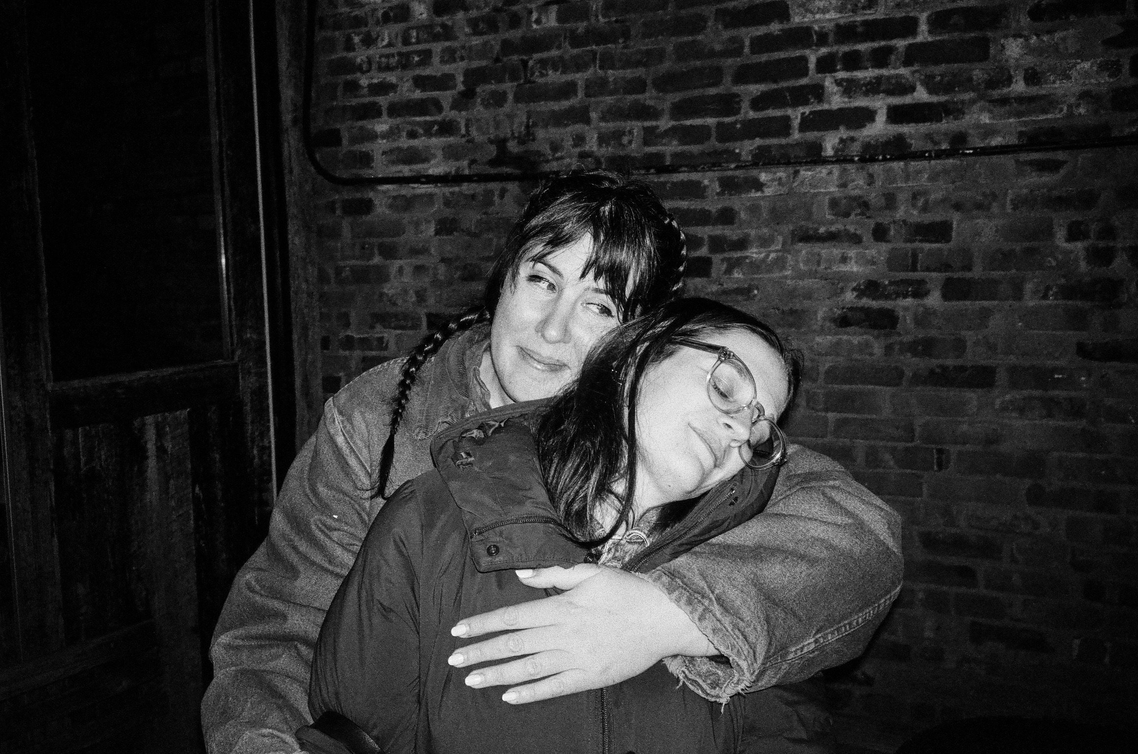 Two women hugging, one with glasses and the other with short hair, in front of a brick wall, black and white photograph.