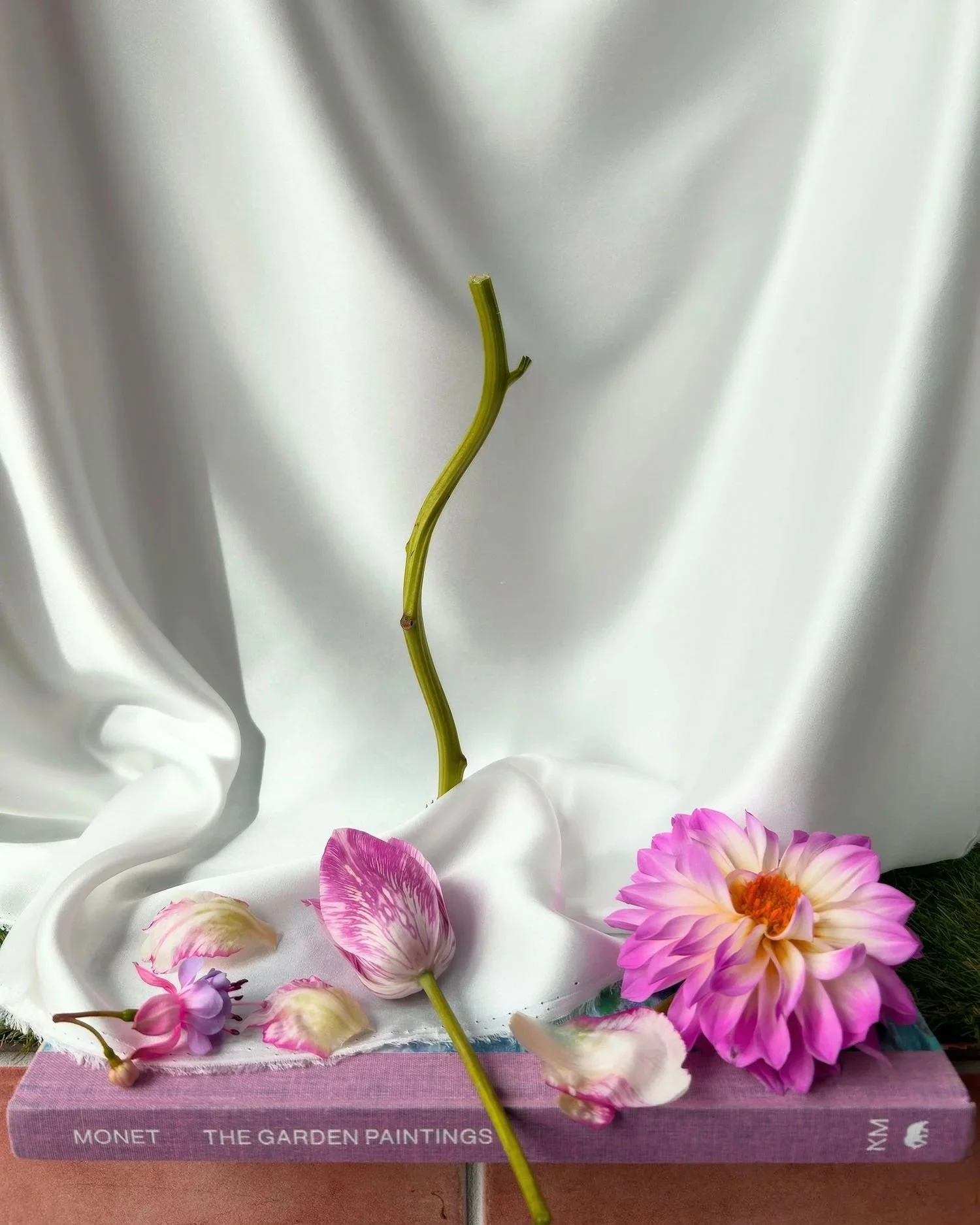 Arrangement of pink and white flowers on top of a purple book titled 'Monet The Garden Paintings' with a white satin fabric backdrop.