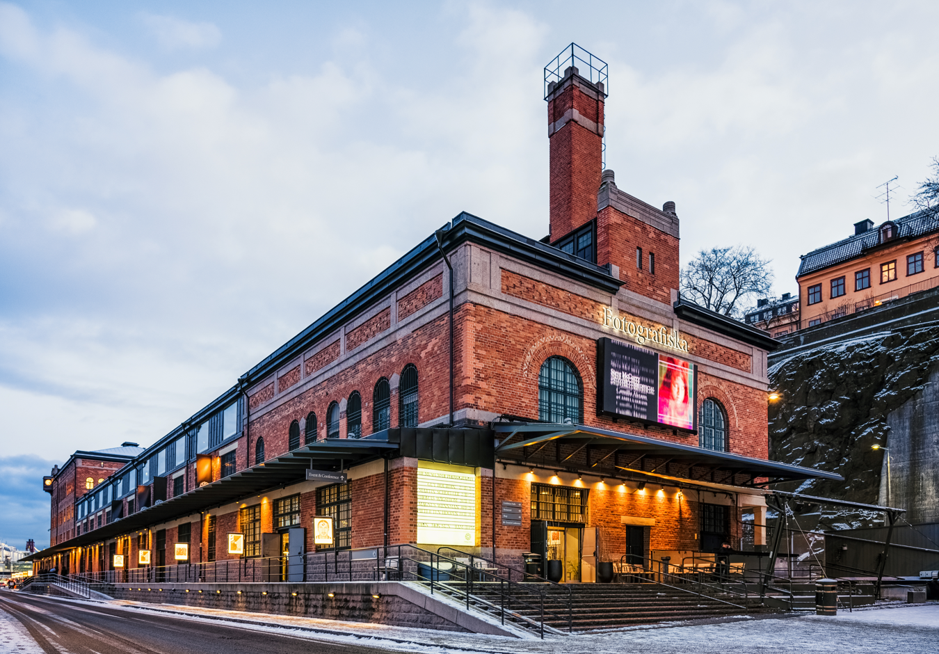 A brick building with illuminated signs and large windows, located on a street with snow and ice, and a cloudy sky overhead.