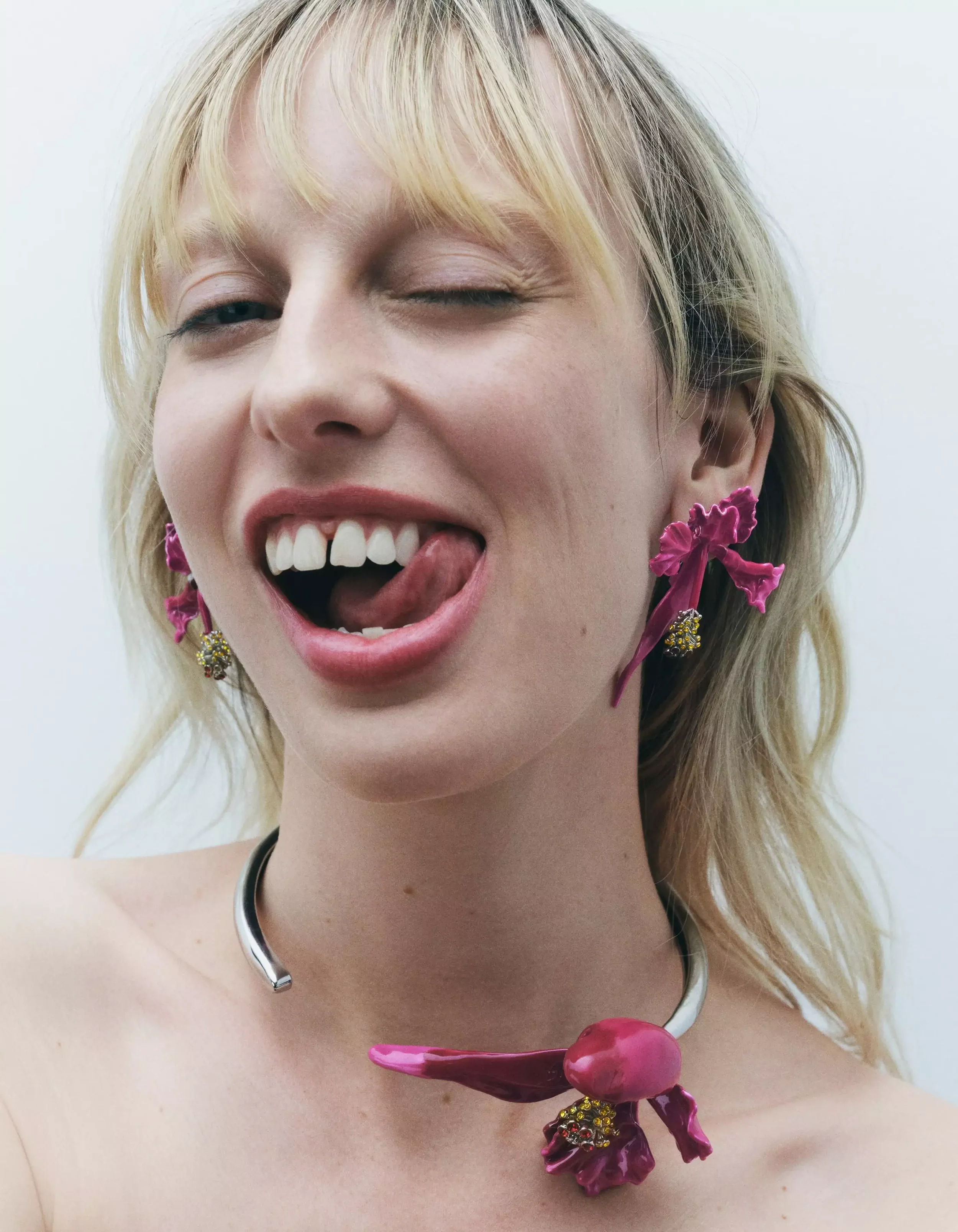 Close-up of a woman with blonde hair, winking and sticking out her tongue, wearing artistic earrings and a matching necklace with pink floral and fruit designs.