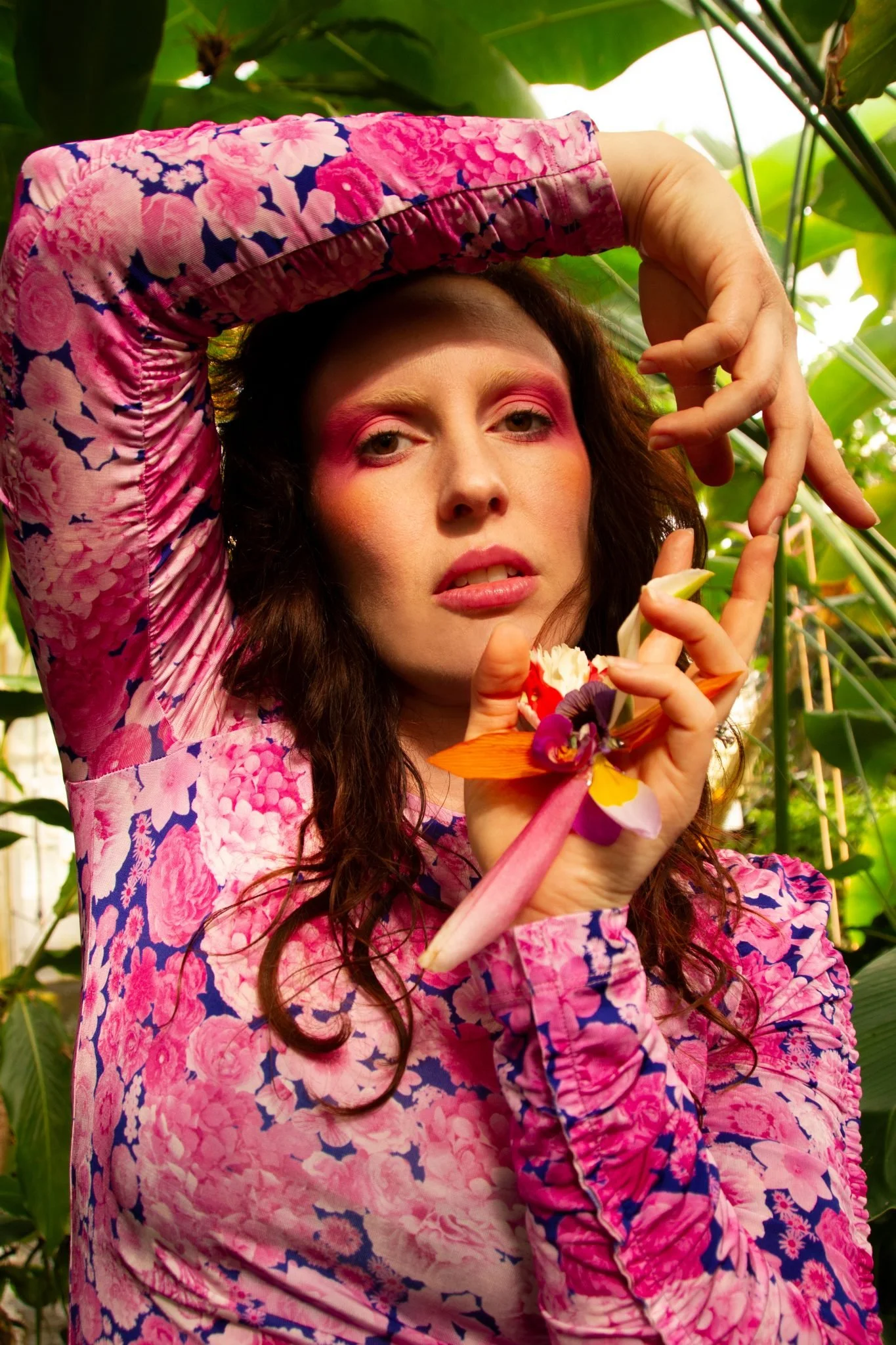 A woman with pink makeup and curly brown hair wearing a pink floral dress, holding a vibrant orchid flower, surrounded by green leaves.