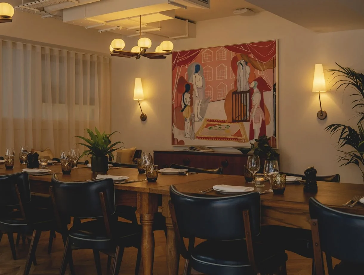 A dining room with a large wooden table set with glasses, plates, napkins, and utensils. There are black chairs around the table, a potted plant as a centerpiece, and a painting on the wall depicting an abstract scene with human figures. The room has warm lighting from wall sconces and a ceiling fixture, with sheer curtains covering windows.