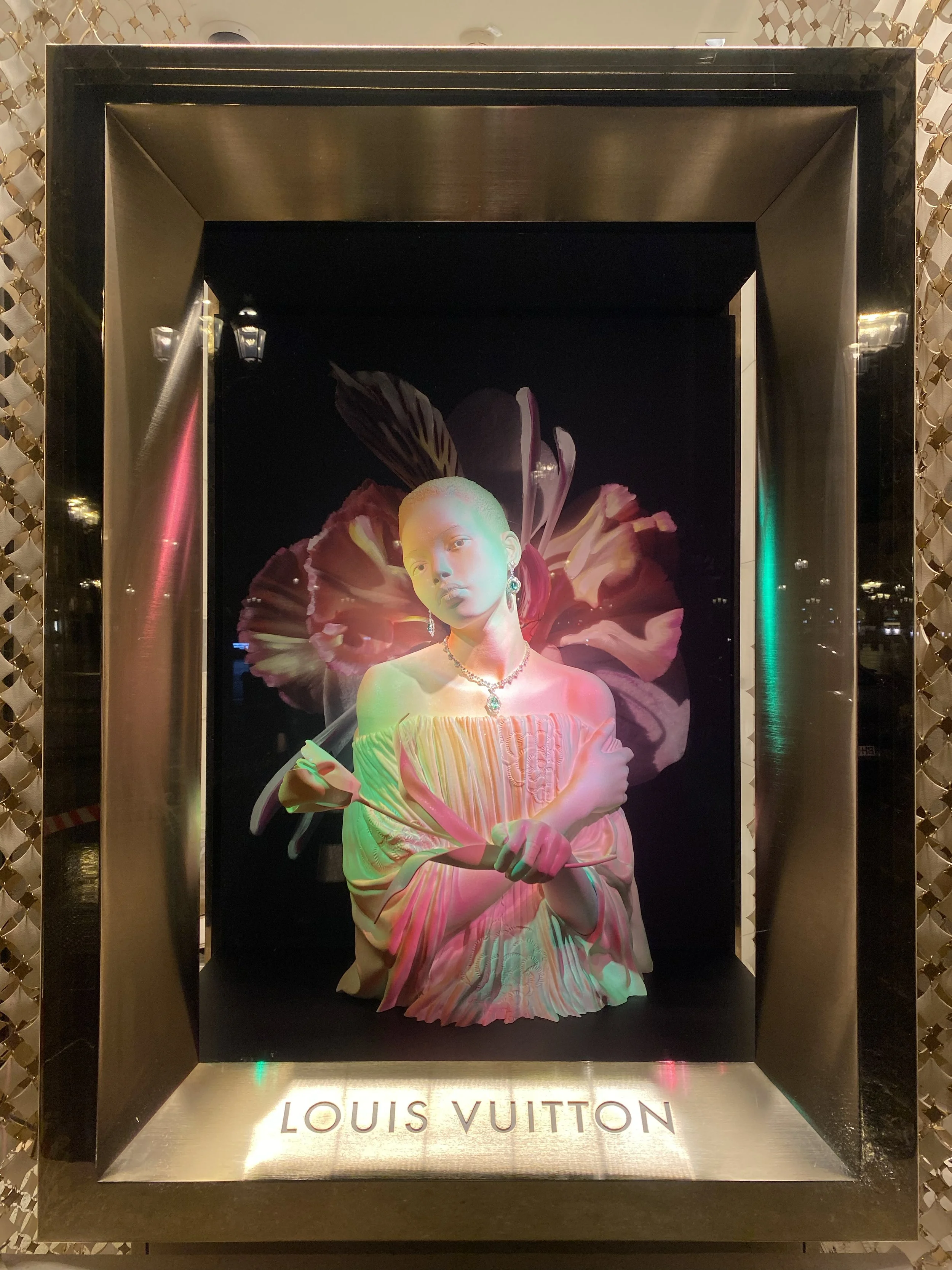 A fashion display window featuring a white mannequin dressed in an off-the-shoulder blouse, with jewelry, and surrounded by floral and feather motifs, with the words "LOUIS VUITTON" at the bottom.