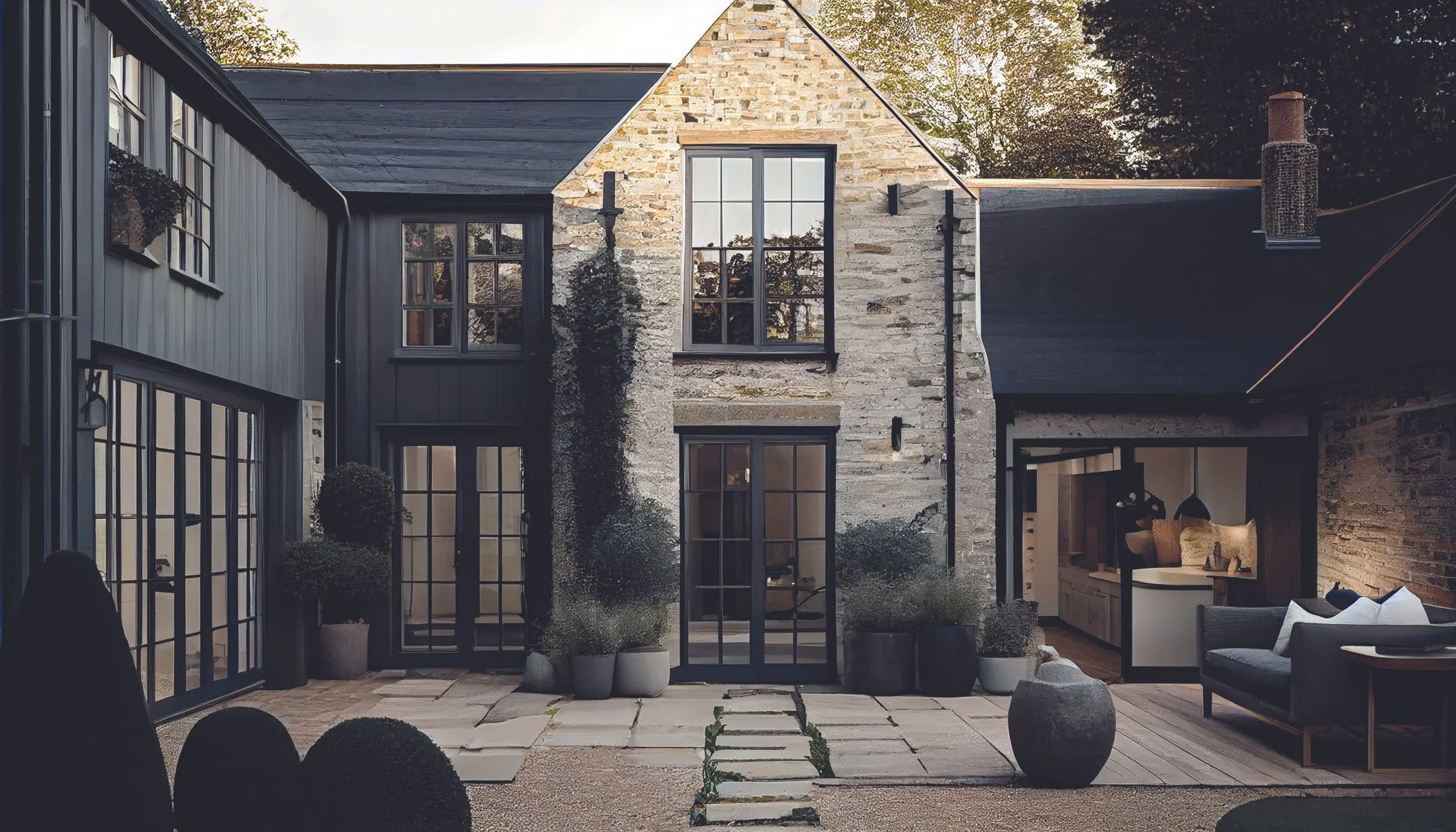 A modern house with a stone facade, large black-framed windows, and a black roof. The outdoor patio has potted plants, patio furniture, and a pathway made of stepping stones.