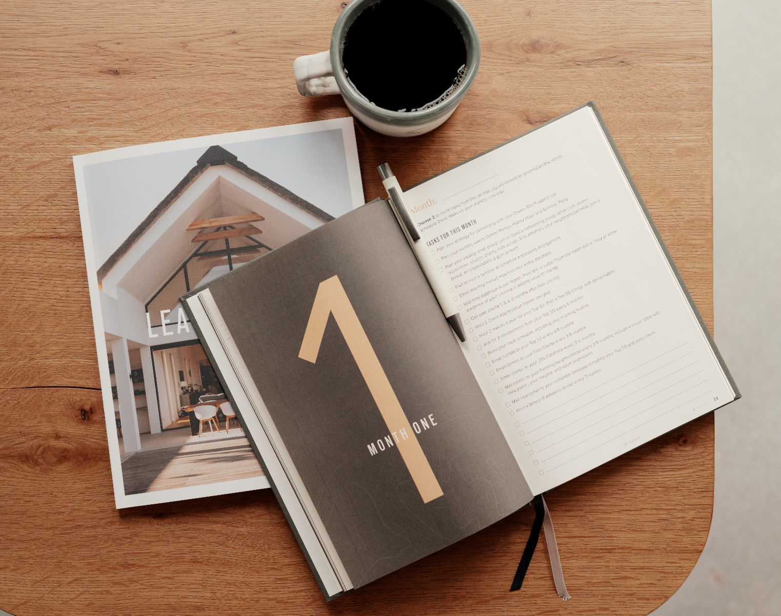 Open planner on wooden table with a black pen, a cup of black coffee, and a pamphlet showing a house with an outdoor seating area.