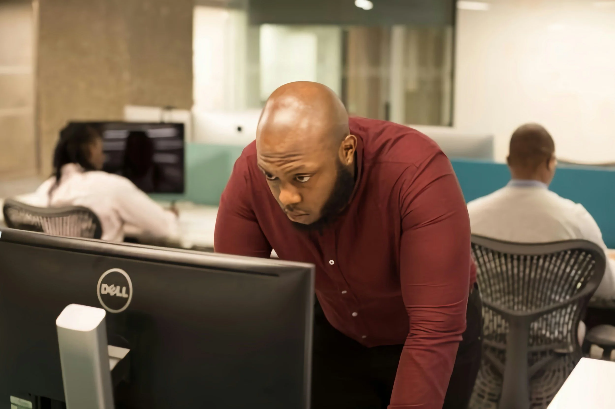 A man with a shaved head and beard leaning over a desk in an office, focused on a computer monitor. Two other people are working at desks in the background.