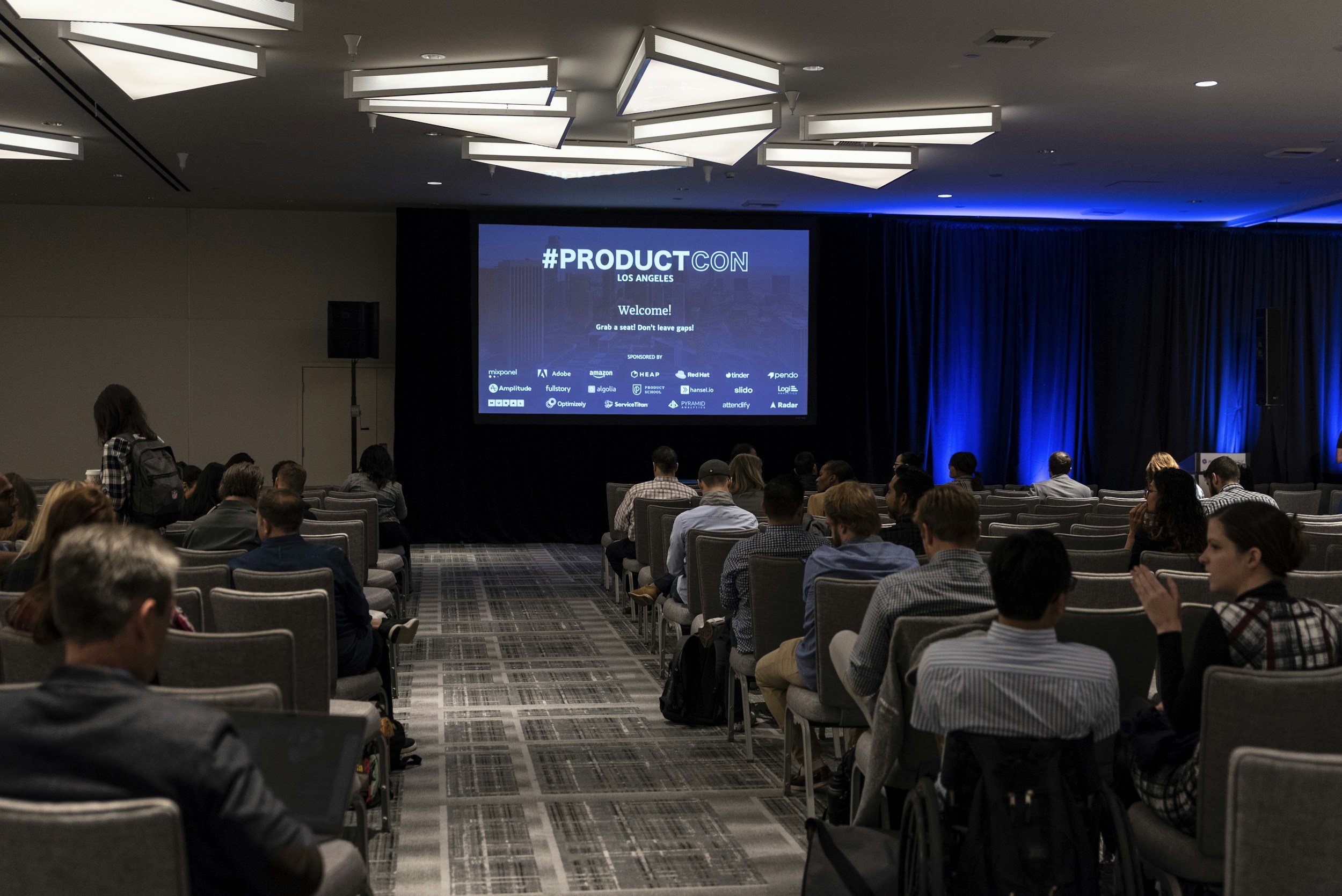 Conference room filled with people watching a presentation on a large screen at #ProductCon Los Angeles, with dim lighting and blue accent lights on the stage.
