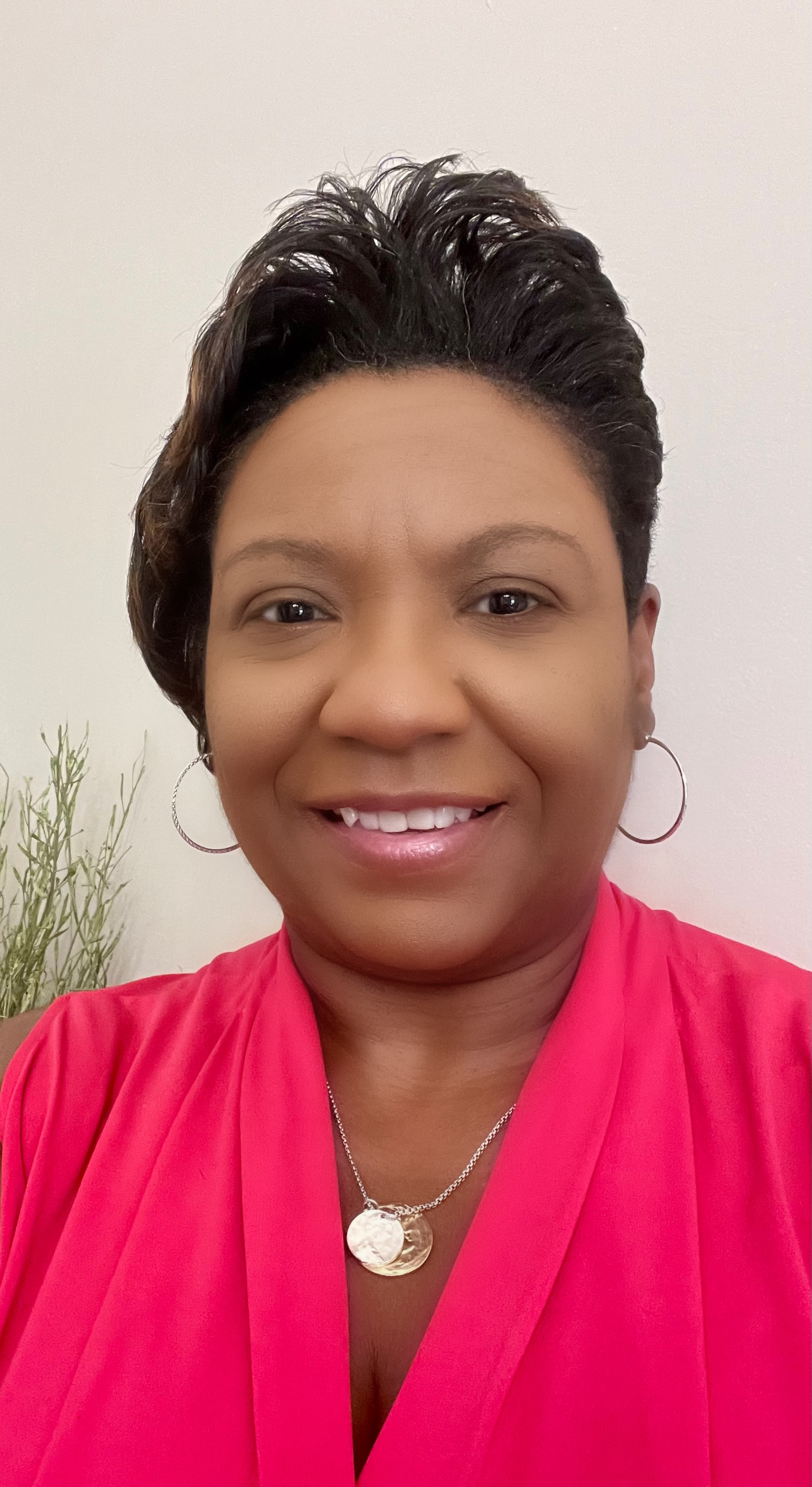 A woman with short, dark, styled hair wearing a pink top, hoop earrings, and a silver necklace with a round pendant. She is smiling and standing in front of a plain white wall with some plant foliage visible on the left side.