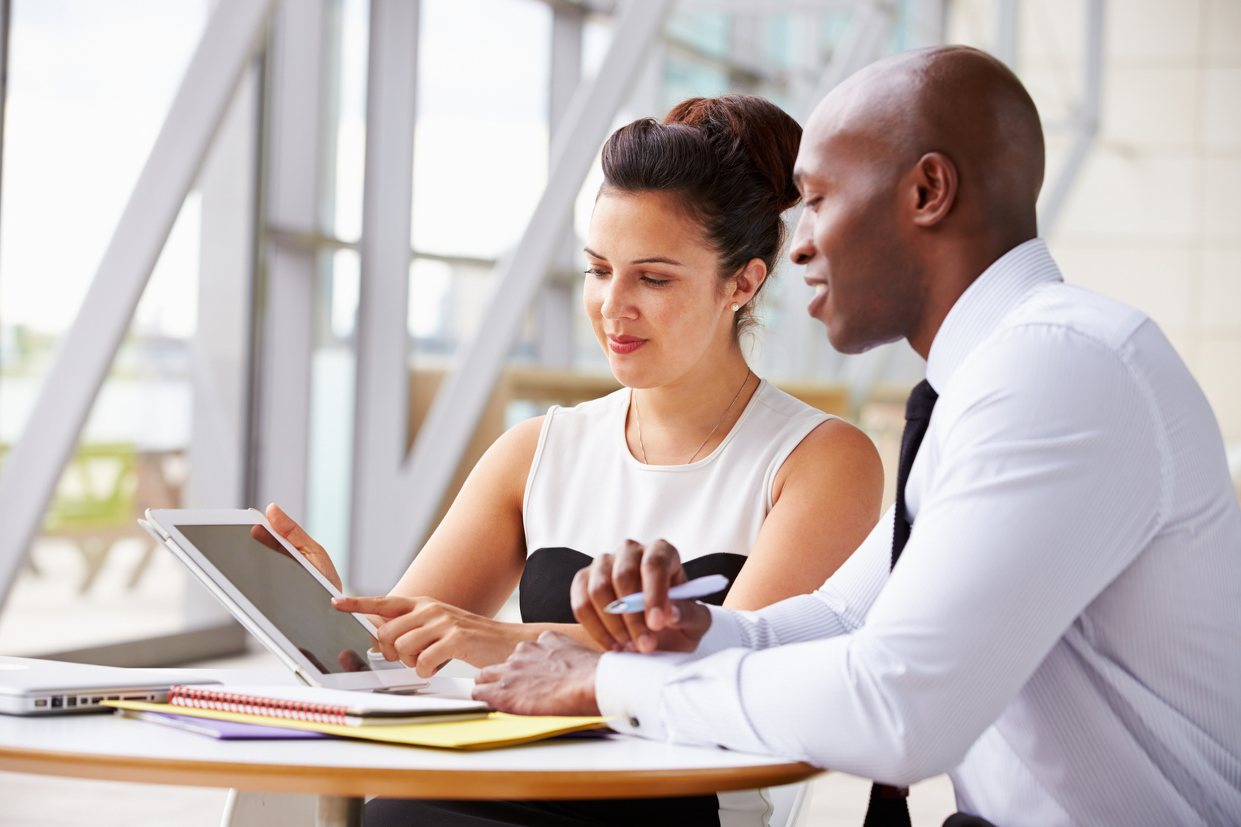 A man and woman in professional attire sitting at a desk in a modern office, discussing something on a tablet device.