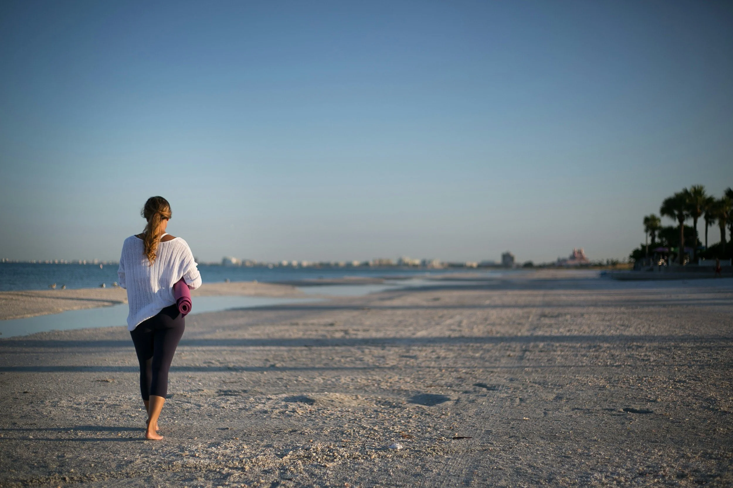 A woman walking barefoot on a sandy beach with a rolled-up yoga mat in her hand, under a clear blue sky with some palm trees in the distance.