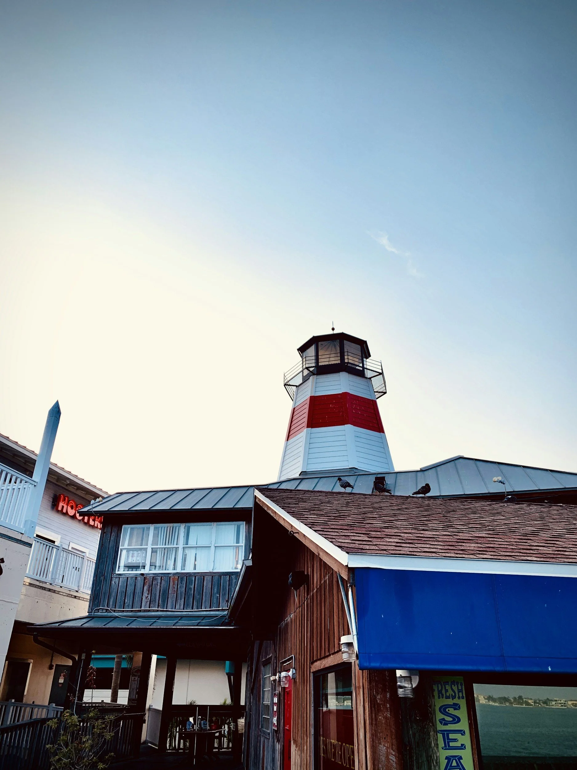 Lighthouse with red and white stripes, wooden building with dark wood siding, blue awning, and a view of water in the background
