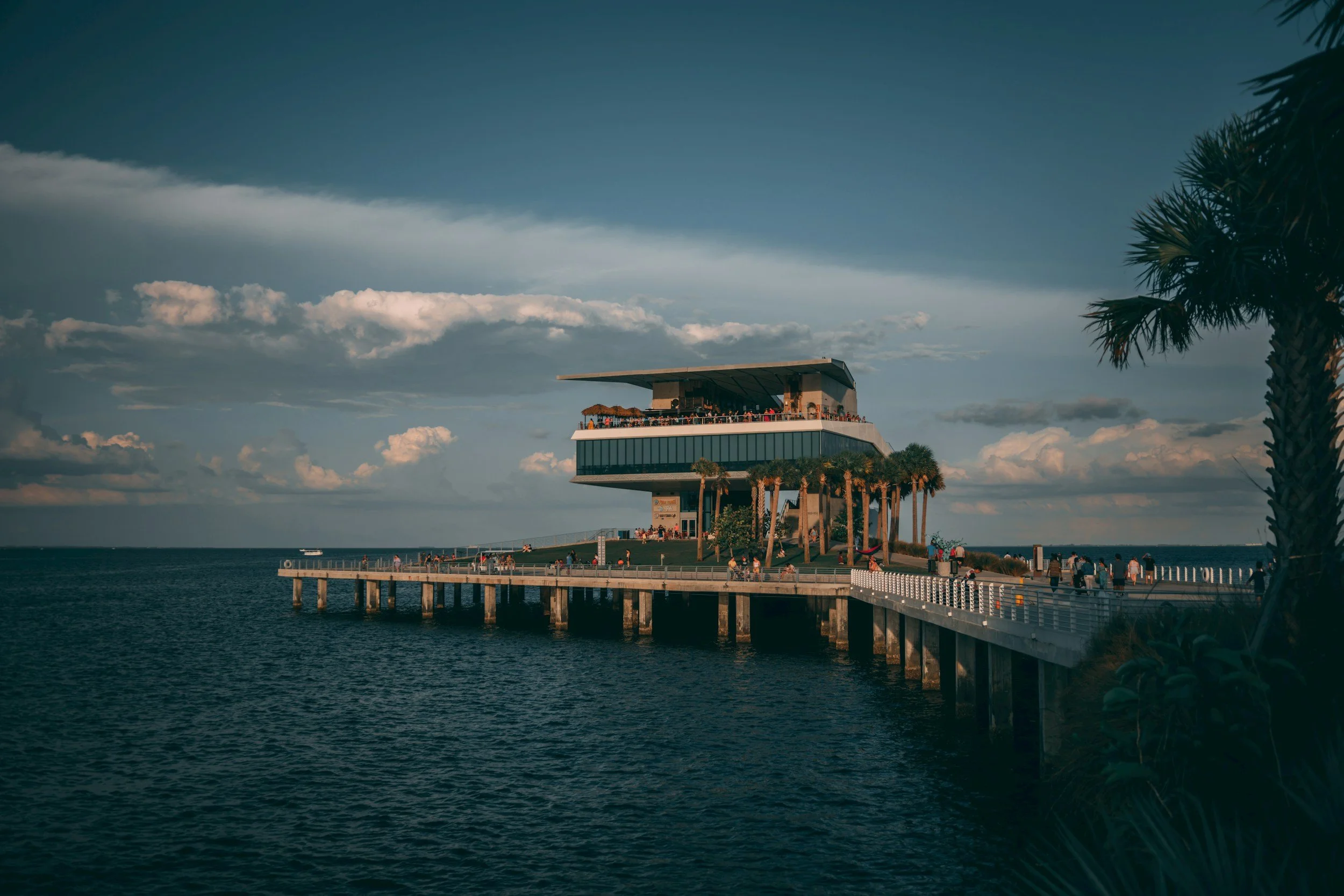 A pier with people going to a modern, elevated building by the water, with palm trees nearby and a cloudy sky
